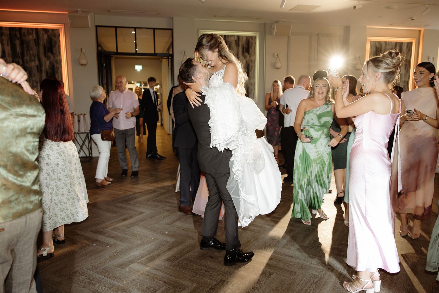 Wedding guests dance and socialize on a dance floor at an indoor reception venue during a celebration.