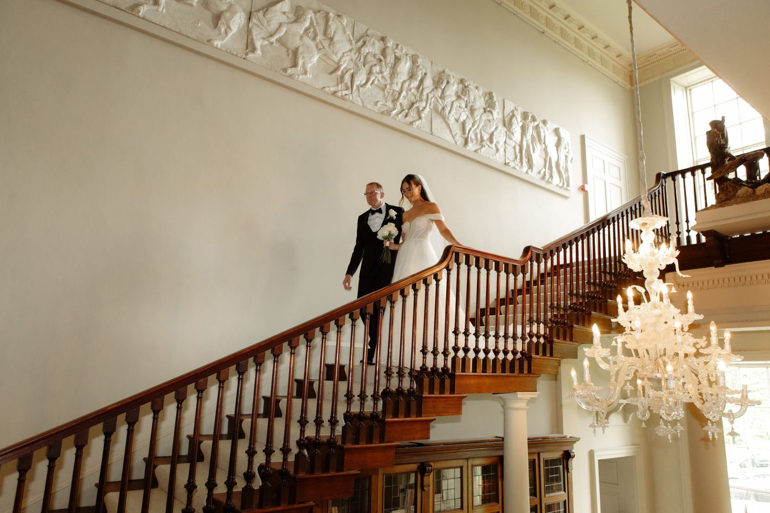 Elegant crystal chandelier hangs in a grand foyer with ornate crown molding and wooden staircase railing.