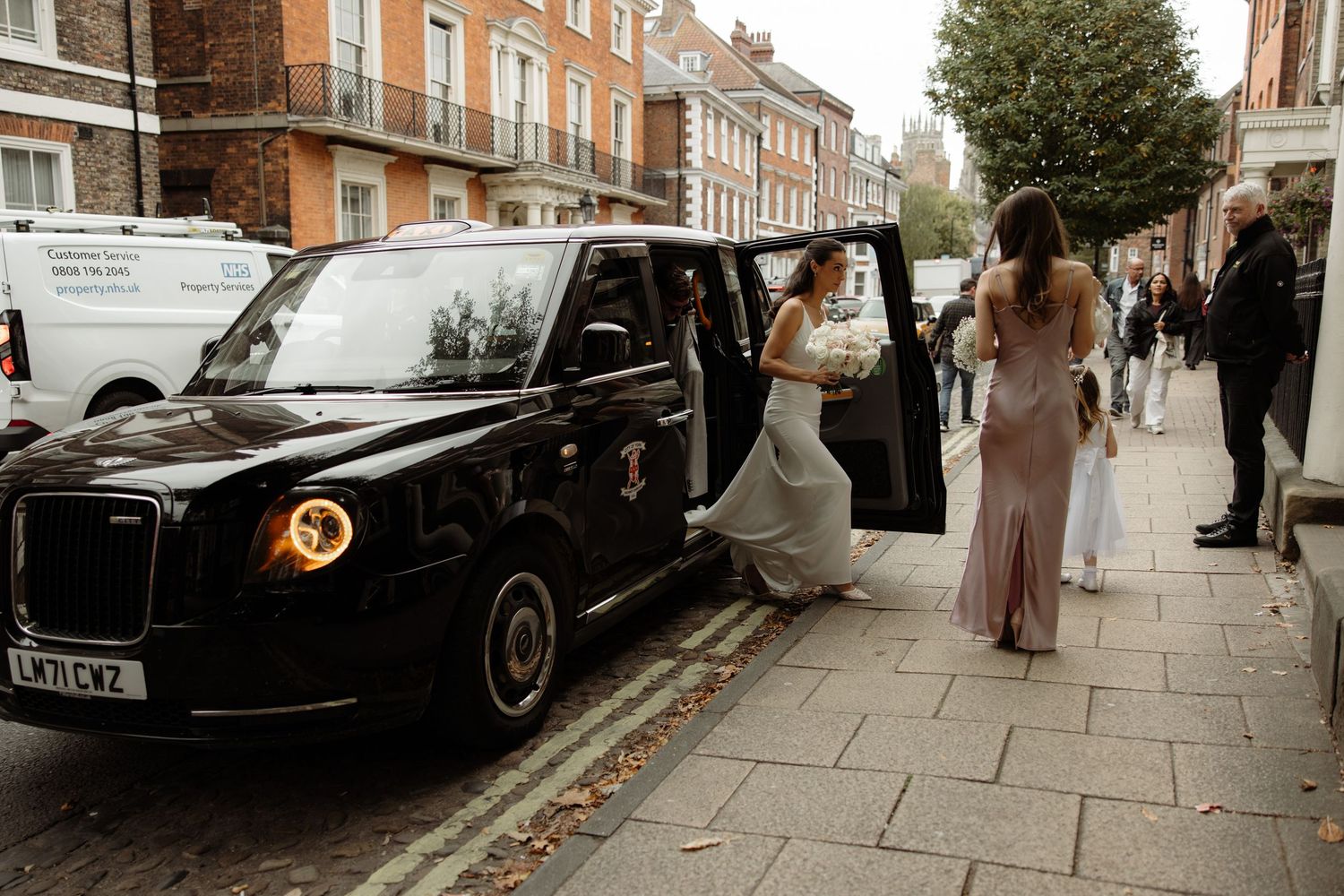 A black London taxi cab stops on a sidewalk as wedding guests in formal attire gather around the vehicle.