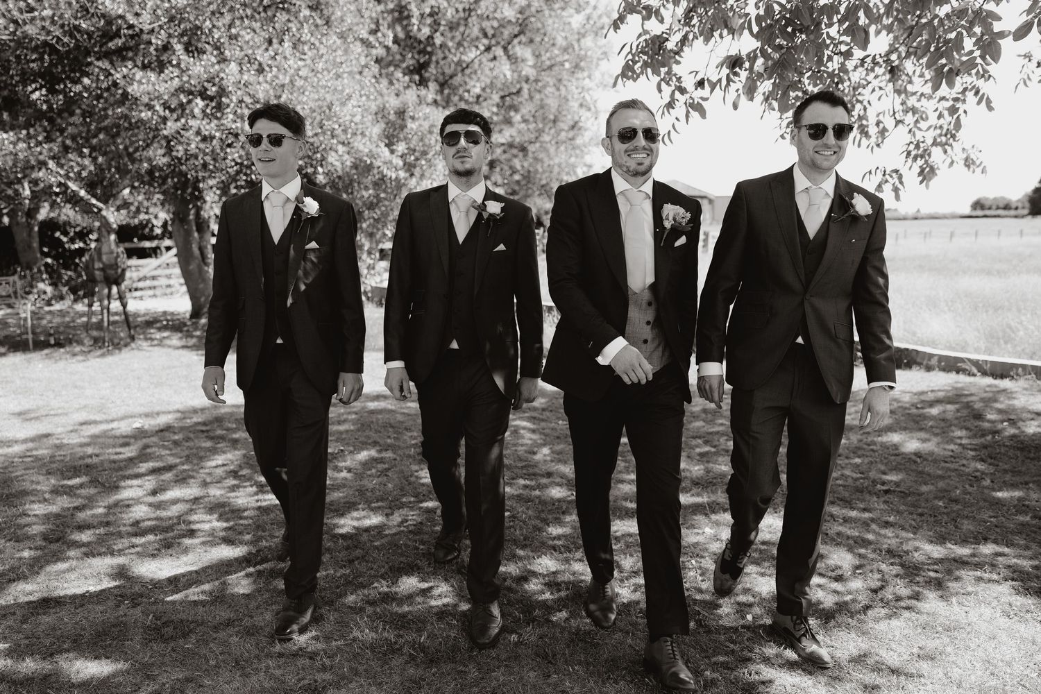 Groomsmen in black suits and sunglasses pose together outdoors under tree shade.