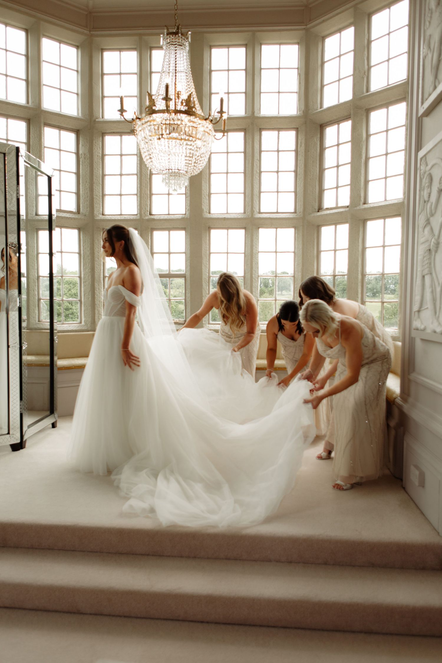 A stunning wedding dress fitting moment in elegant room with large windows and crystal chandelier.