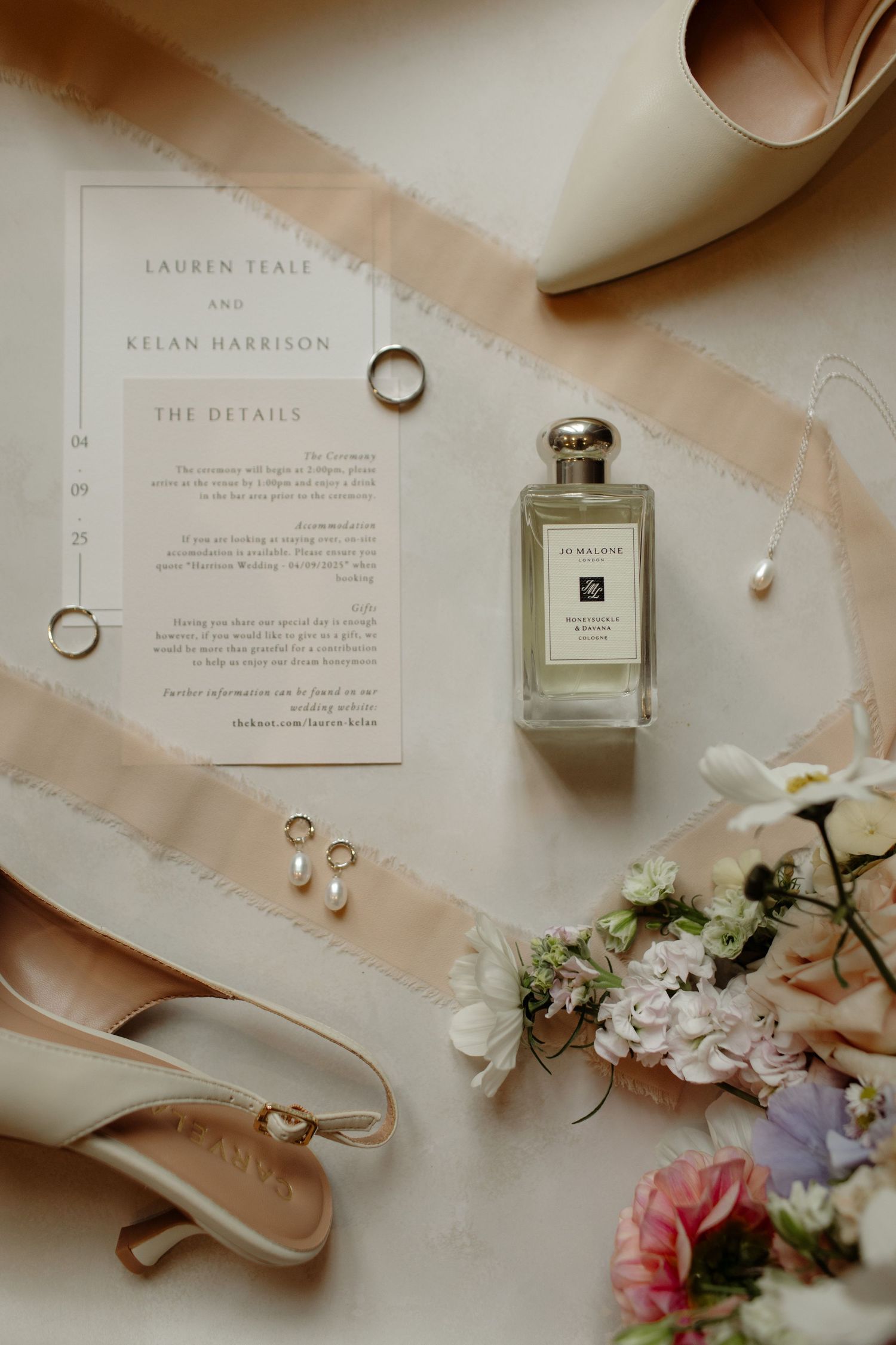 Wedding details flatlay with rings, perfume bottle, invitation card, and delicate floral elements on light background.