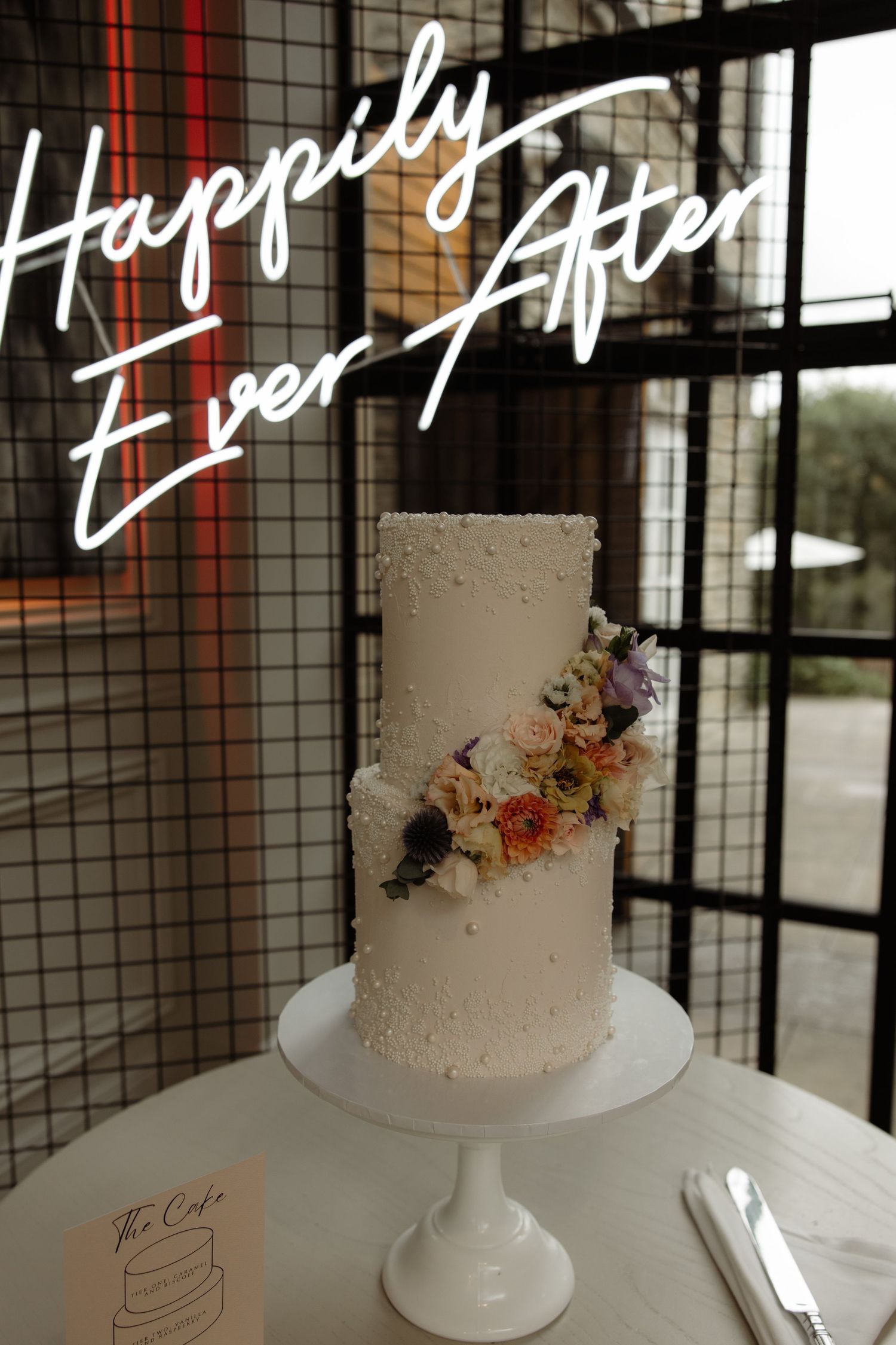Minimalist white wedding cake with delicate floral accents displayed on a pedestal against a dark background.