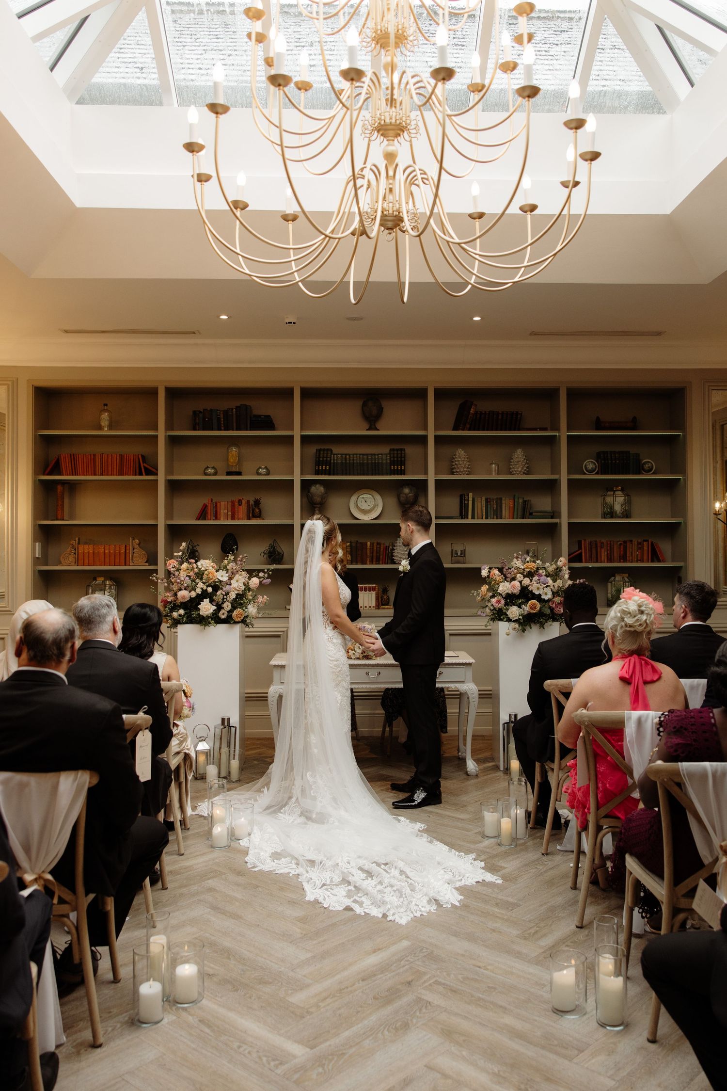 Indoor wedding ceremony in an elegant library room with herringbone floors and a grand gilded chandelier beneath a skylight.