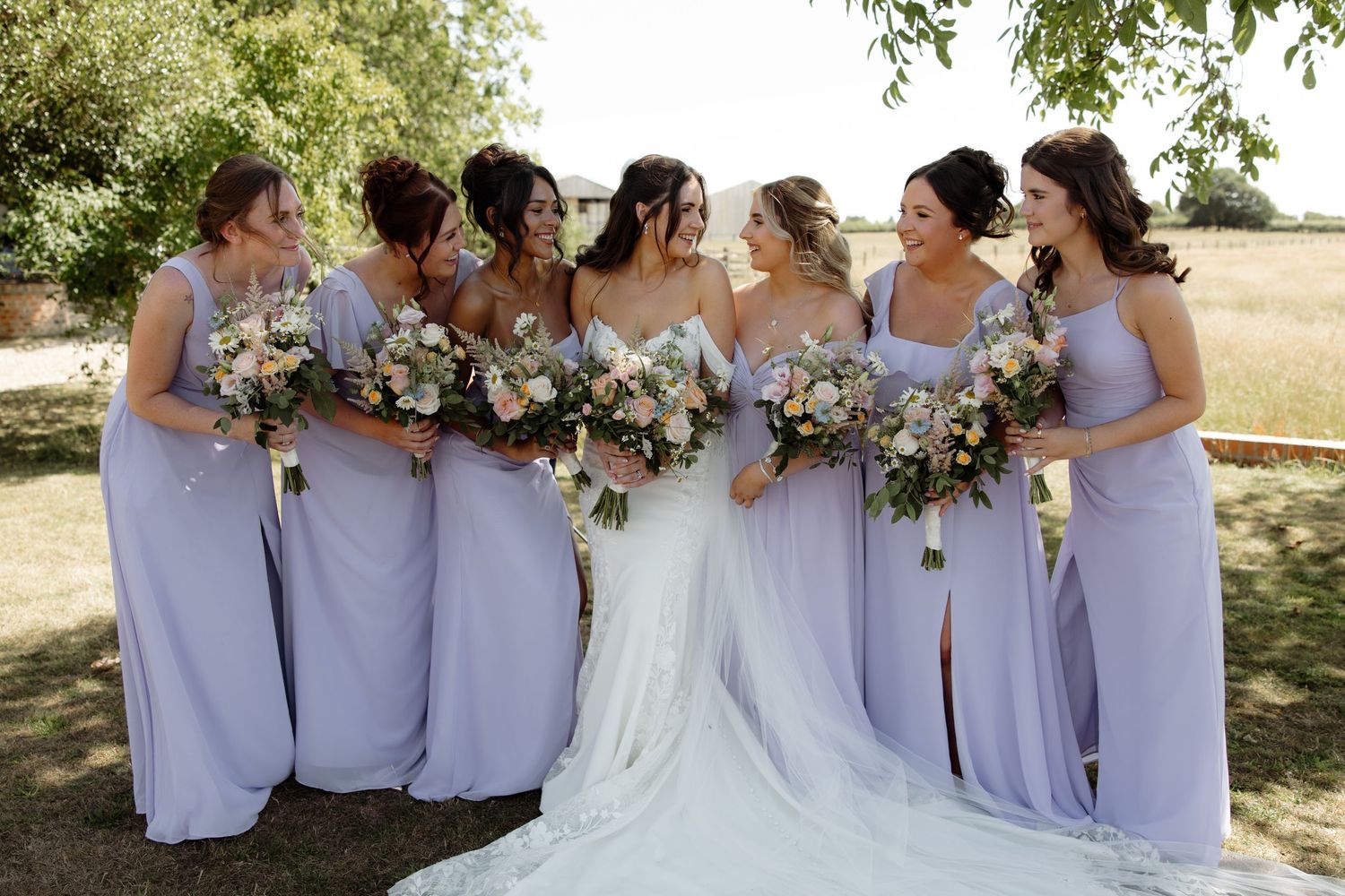 Bridesmaids in lavender dresses pose with bride in white gown outdoors during summer wedding celebration.