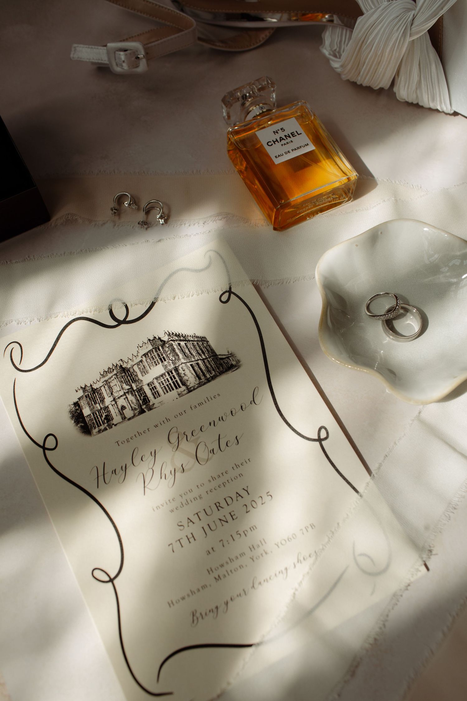 Wedding invitation cards with elegant calligraphy lay scattered among perfume bottles on a white surface in soft lighting.