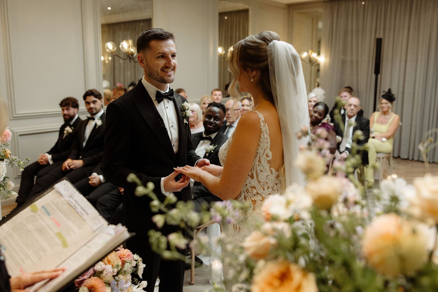 A wedding ceremony in an elegant room with soft lighting and floral arrangements as a couple exchanges vows.