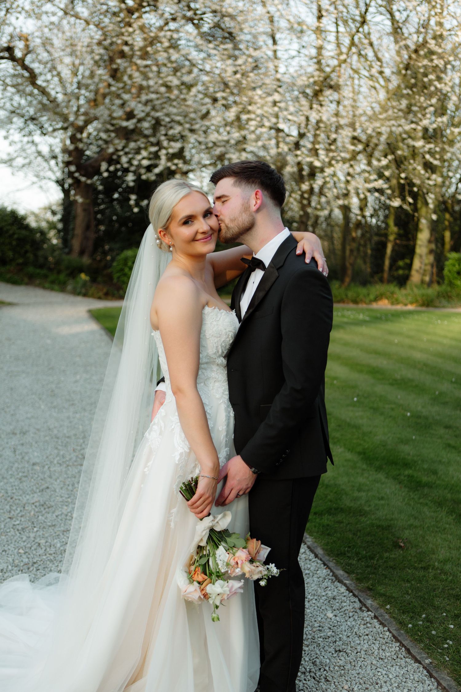 A romantic outdoor wedding portrait showing a couple in wedding attire on a tree-lined path.