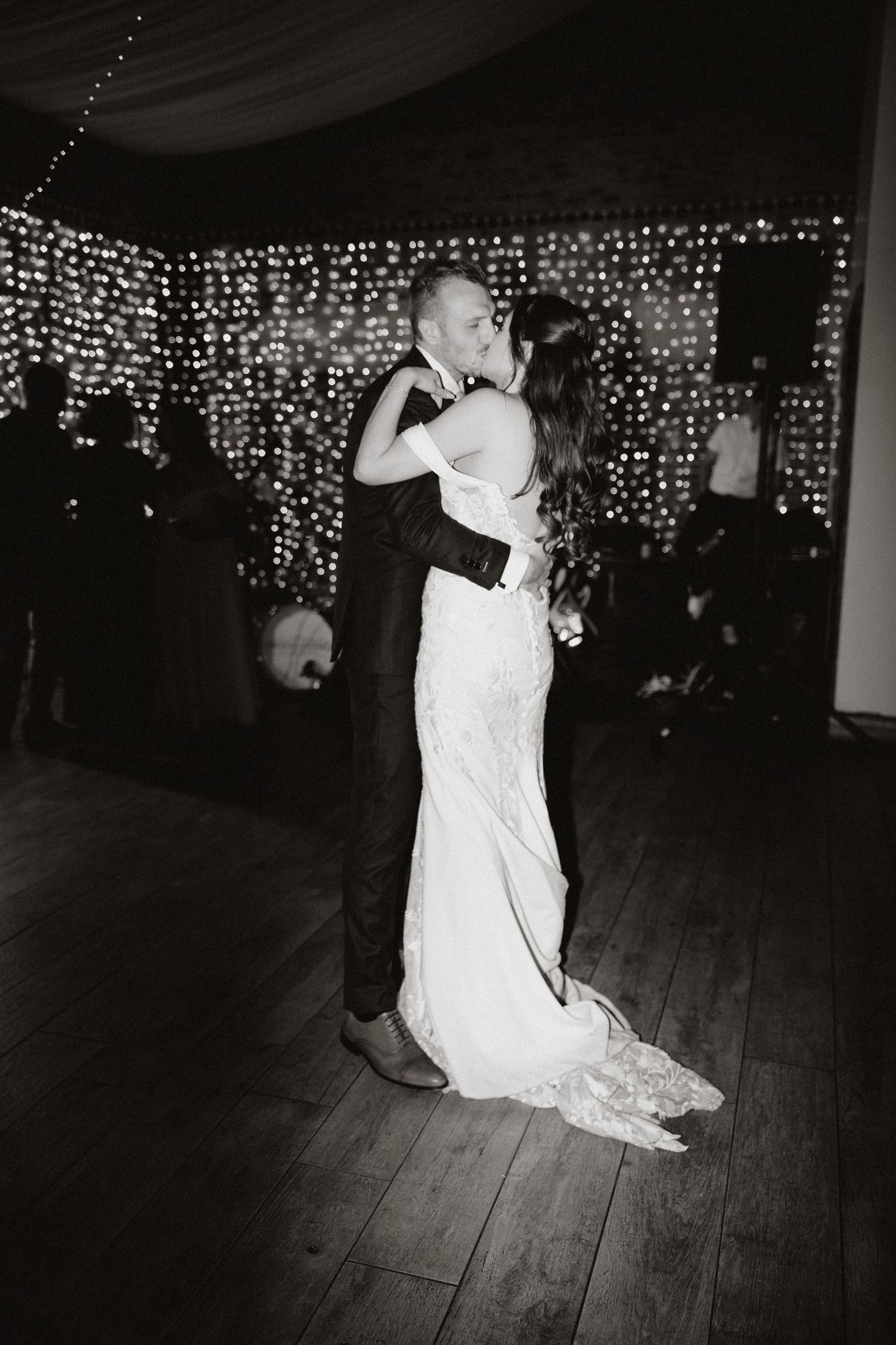 Newlyweds share their first dance under romantic string lights on a wooden dance floor.