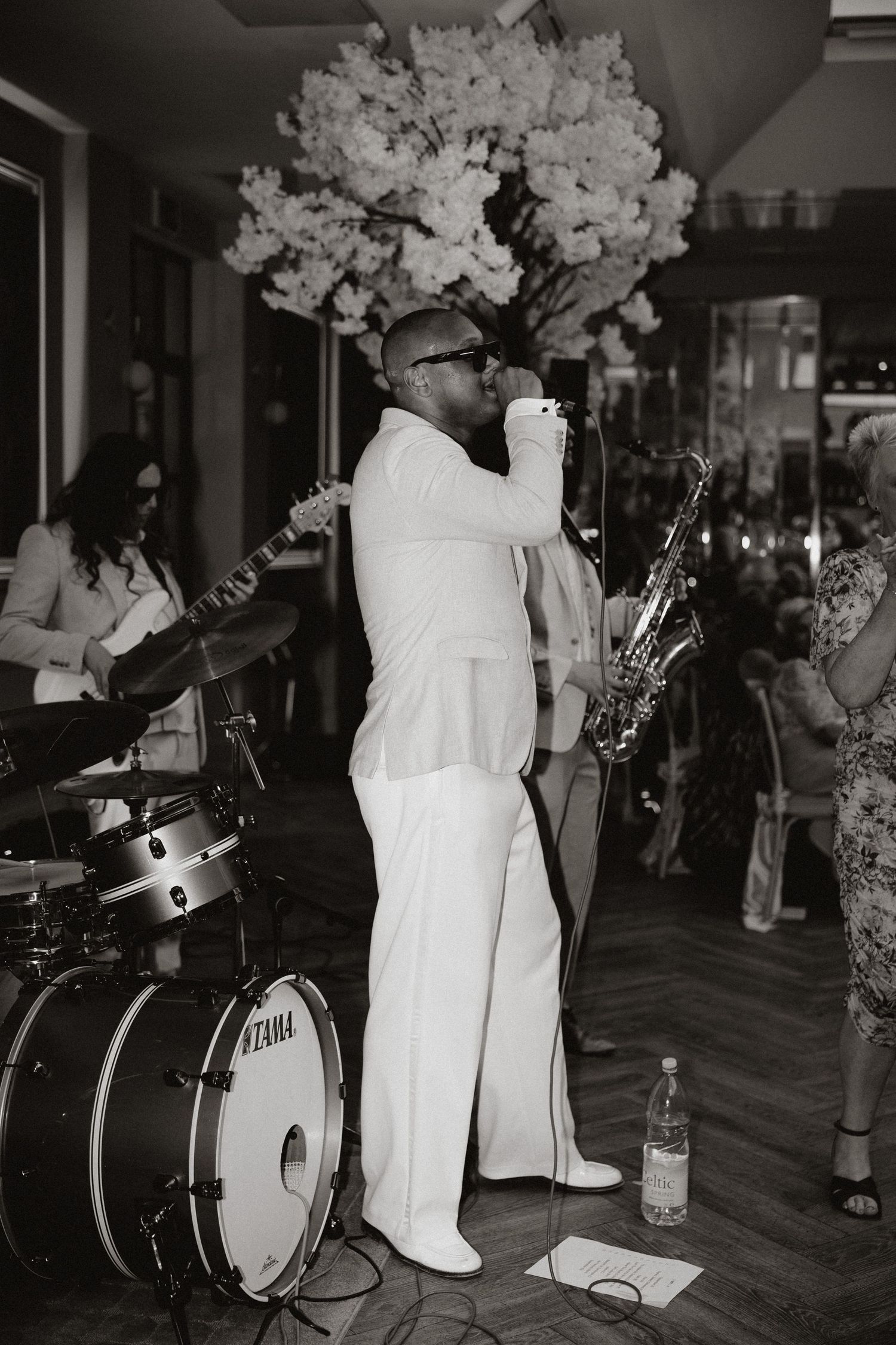 Black and white image of musicians performing at an event with decorative white flower arrangements on stage.