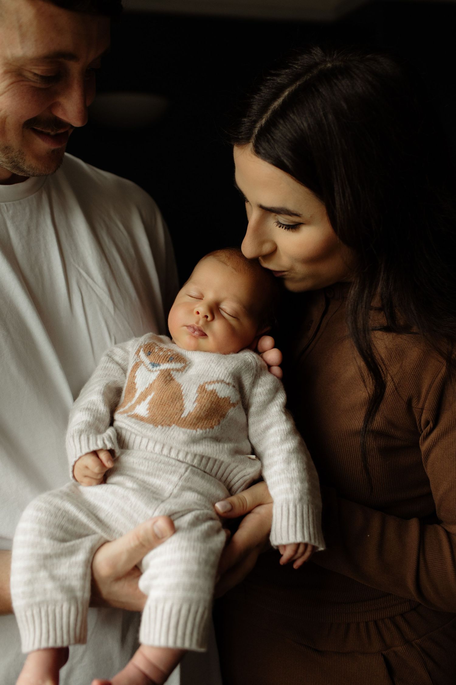 A sleeping baby wearing a white knit sweater is cradled lovingly in moody, dark lighting.