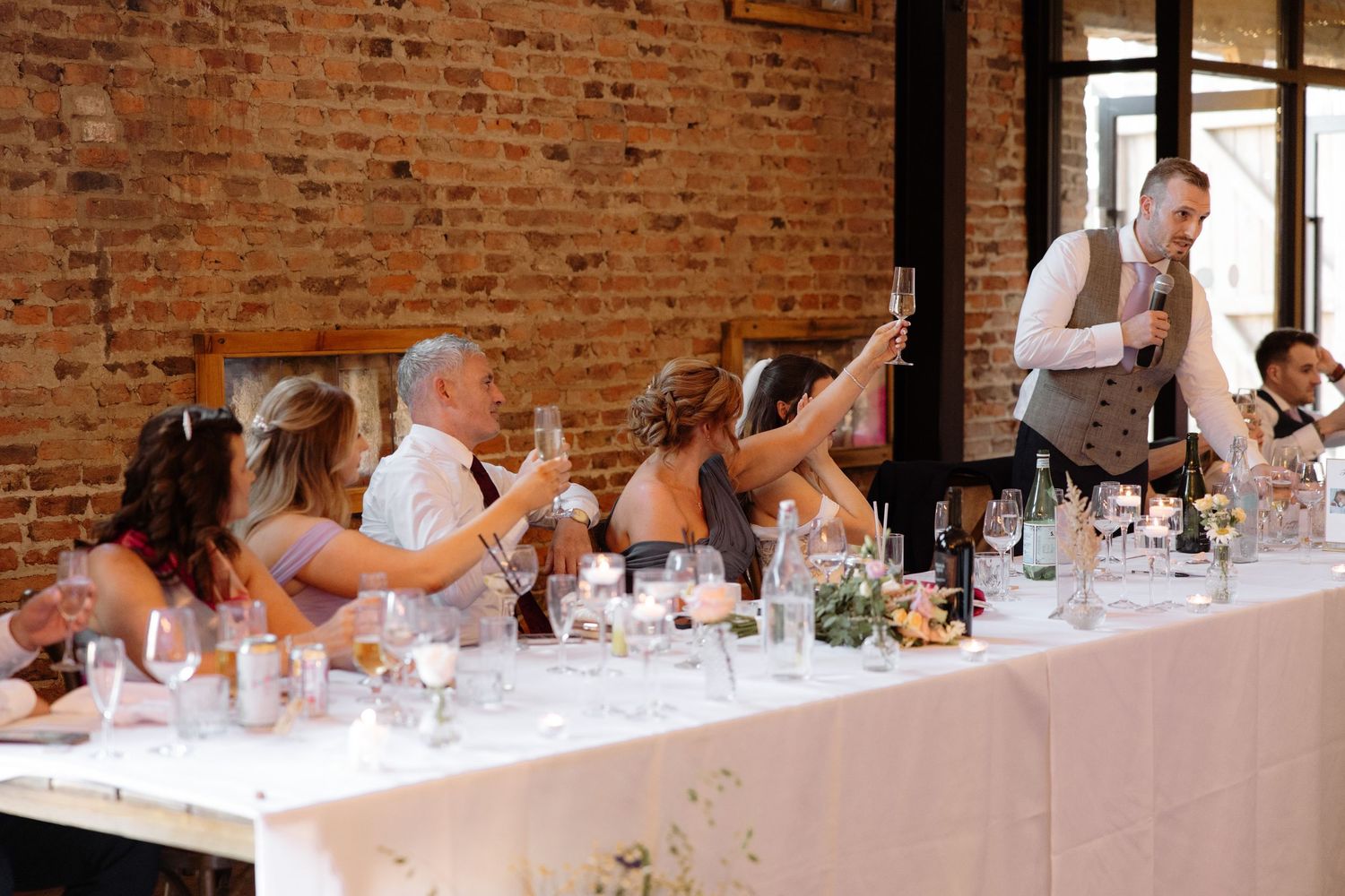 Wedding guests at a long reception table with exposed brick walls react enthusiastically to a toast being given.