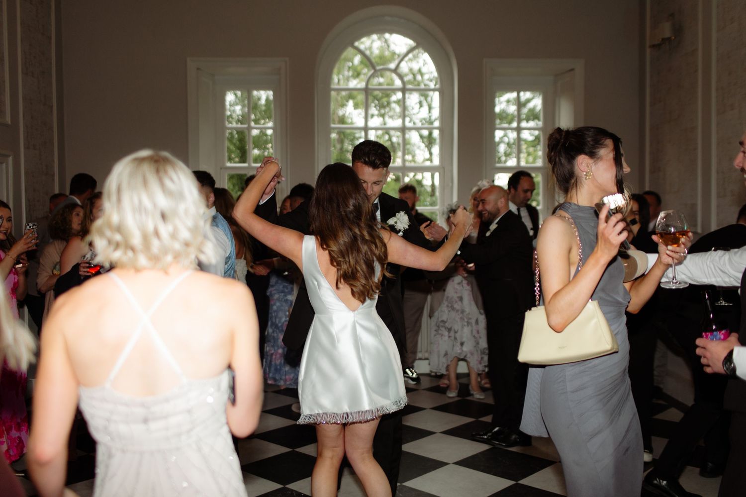 Party guests dancing together at an elegant indoor venue with arched windows and checkered floor.