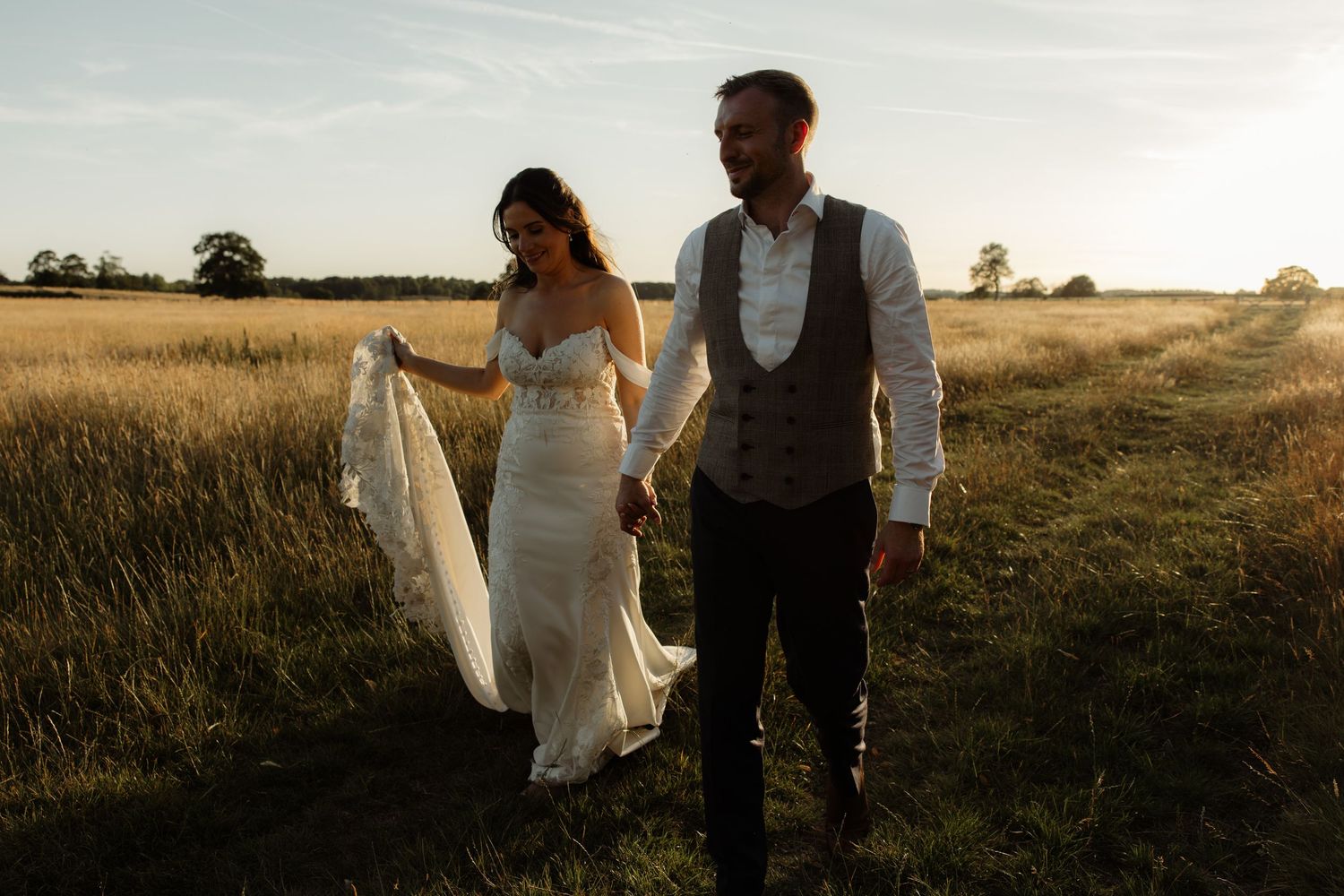 A couple in wedding attire walks together through a golden wheat field at sunset.
