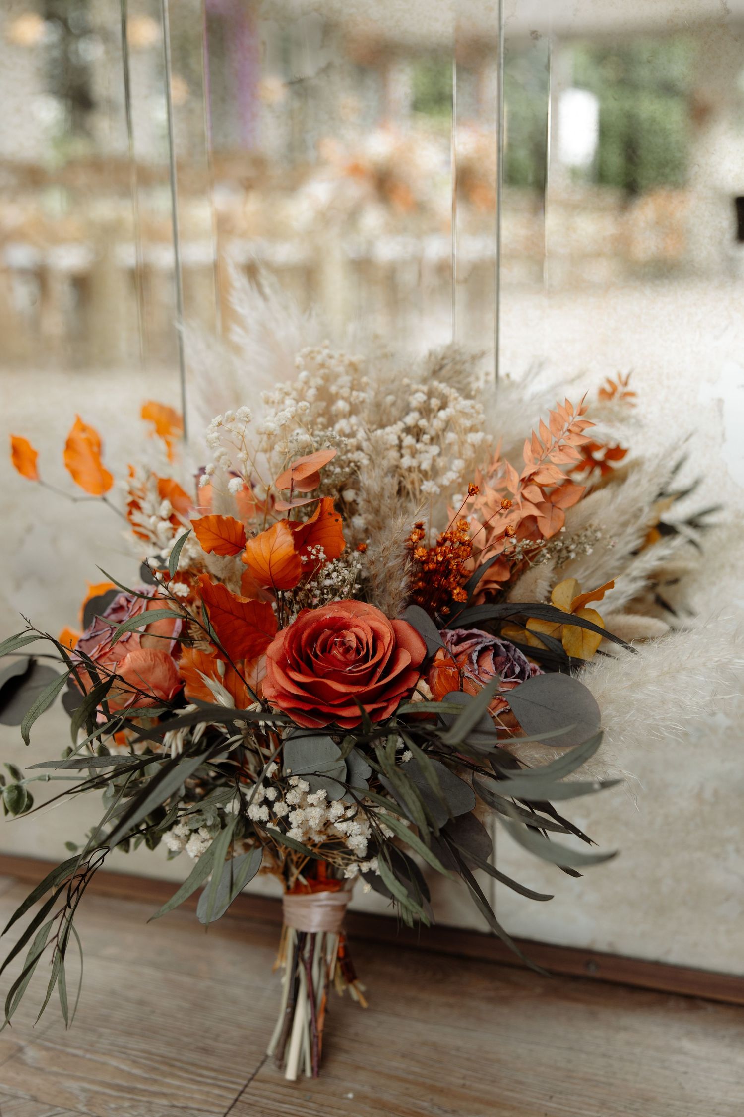 Romantic autumn floral arrangement with orange roses and eucalyptus leaves displayed on rustic wooden table surface.