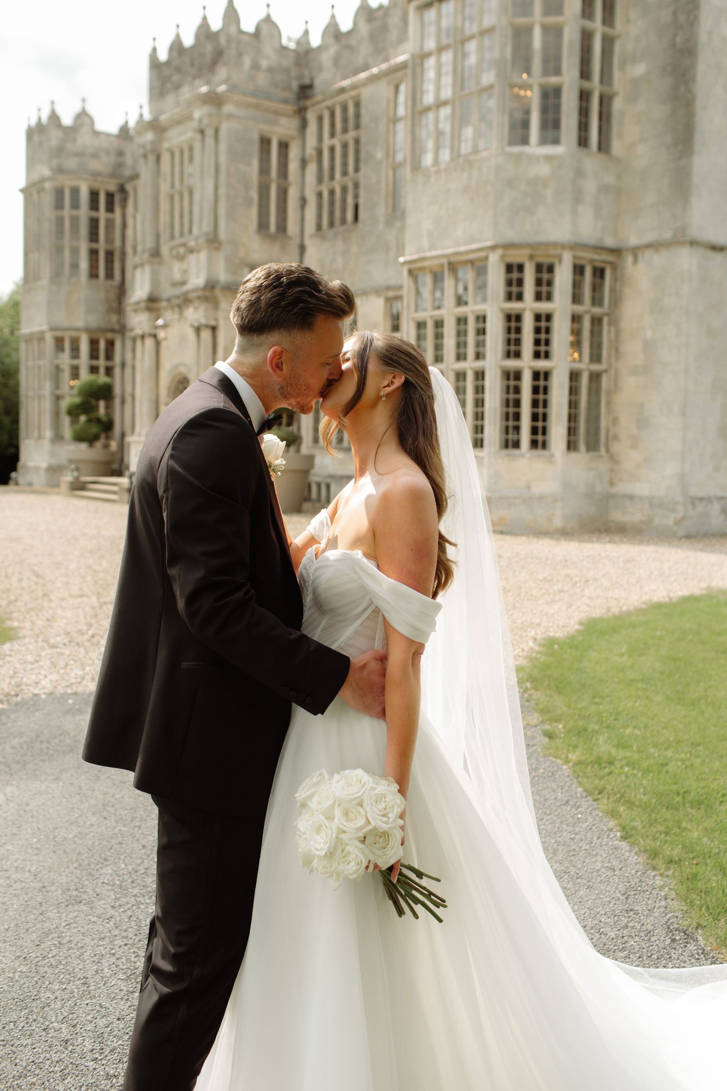 A romantic wedding couple shares a tender moment outside a grand Gothic-style mansion with white bouquet and flowing veil.