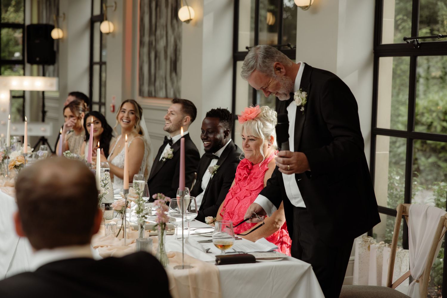 Wedding guests seated at long tables in an elegant reception venue with large windows and chandeliers.