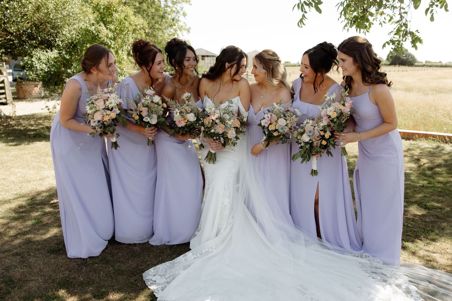 Wedding party in lavender dresses share candid laughing moments together in an outdoor summer setting.