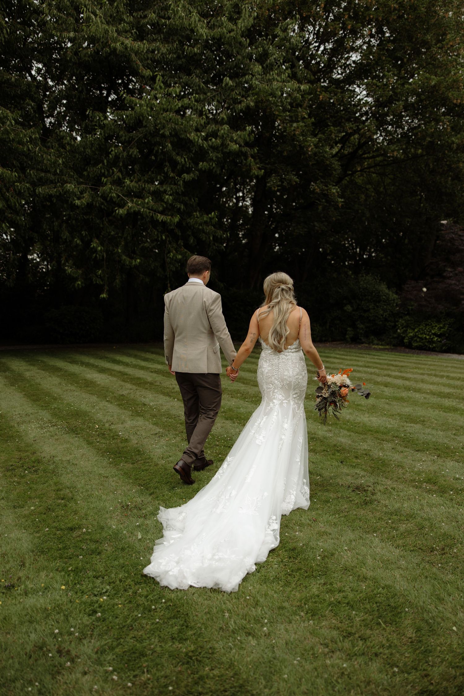 A couple walks away across a lush green lawn, with the bride wearing a flowing white gown and carrying a bouquet.