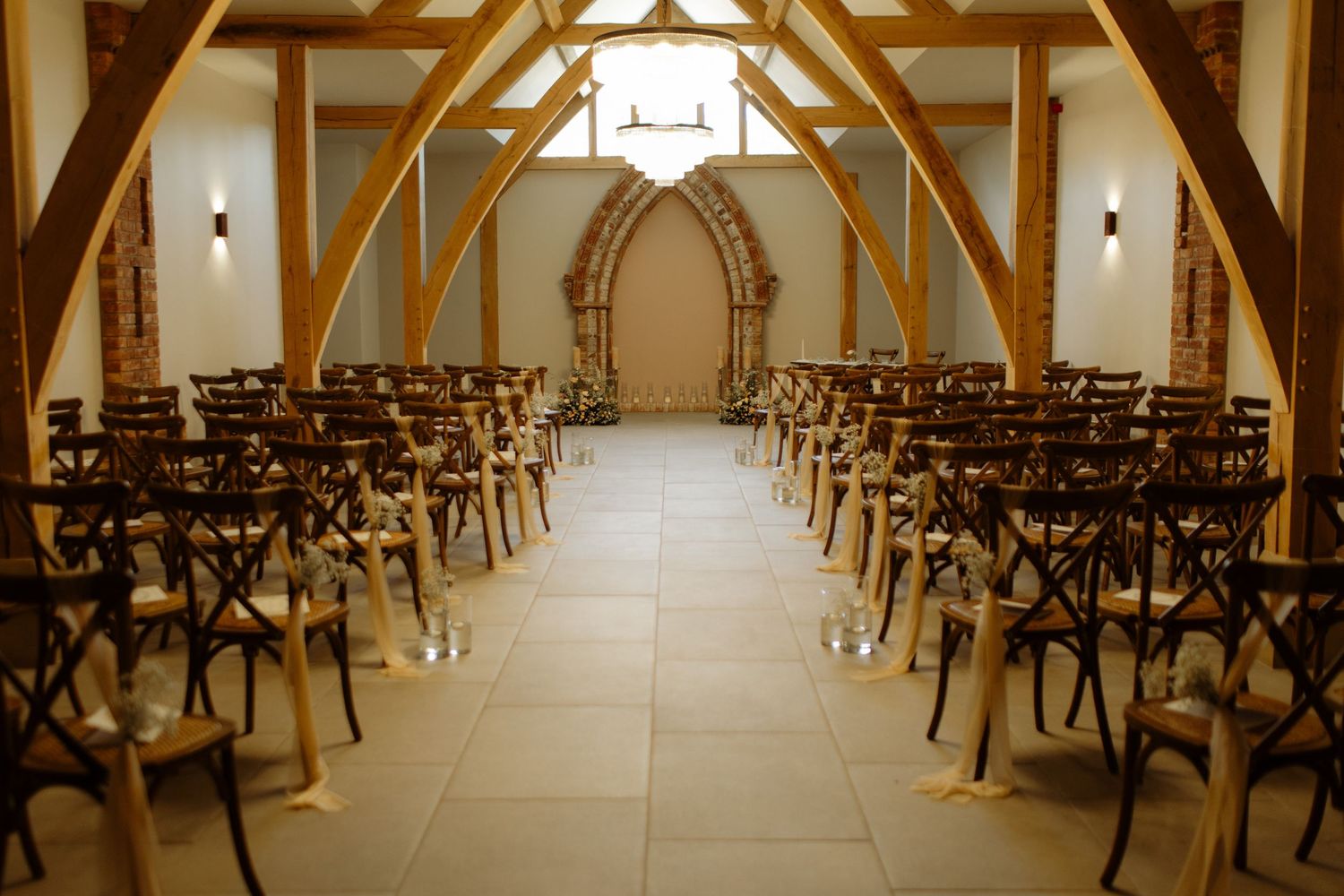 Elegant wedding venue interior with wooden cross-back chairs and exposed wooden beams illuminated by warm lighting.