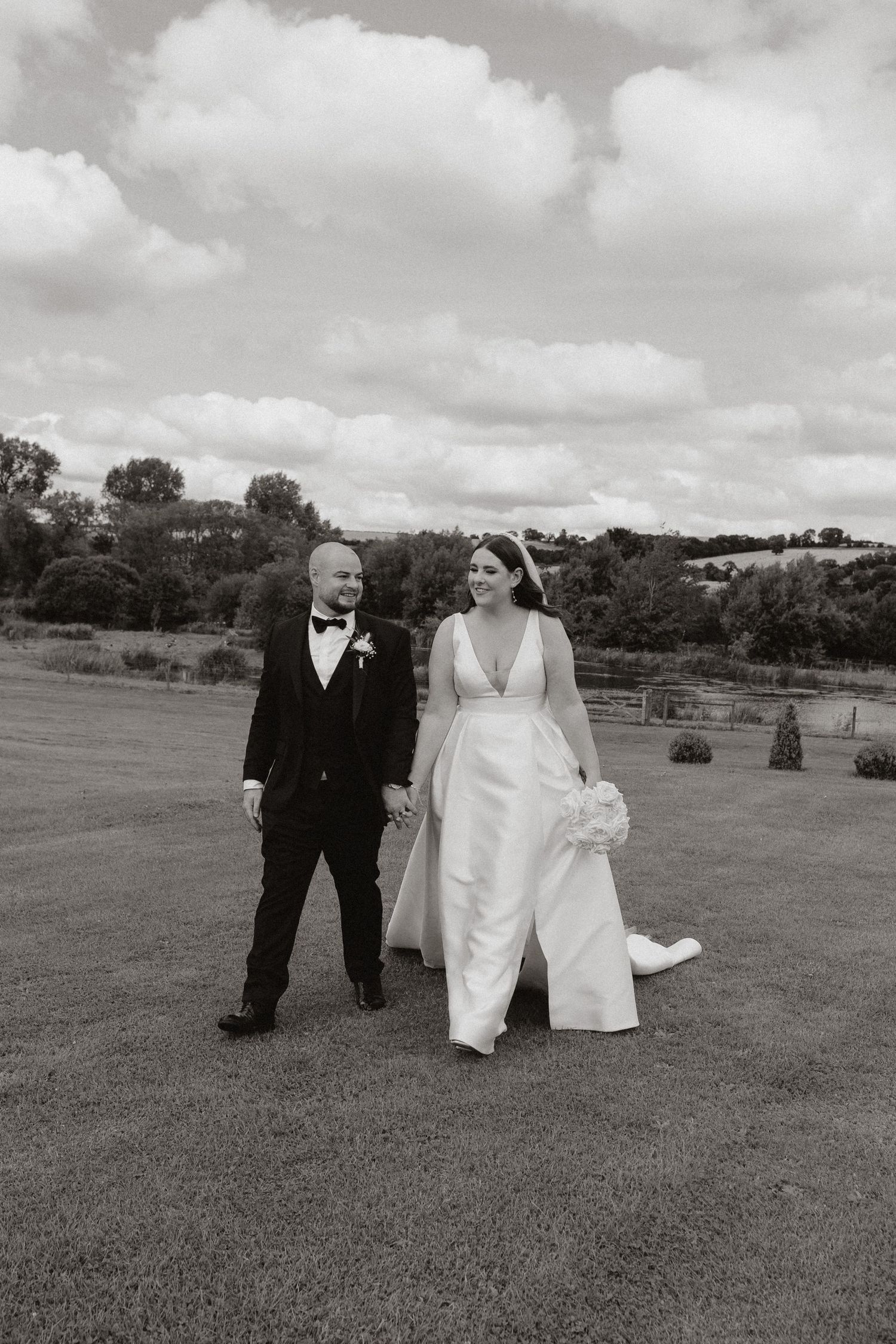 A wedding party walks together across a grassy field on an overcast day, captured in black and white.