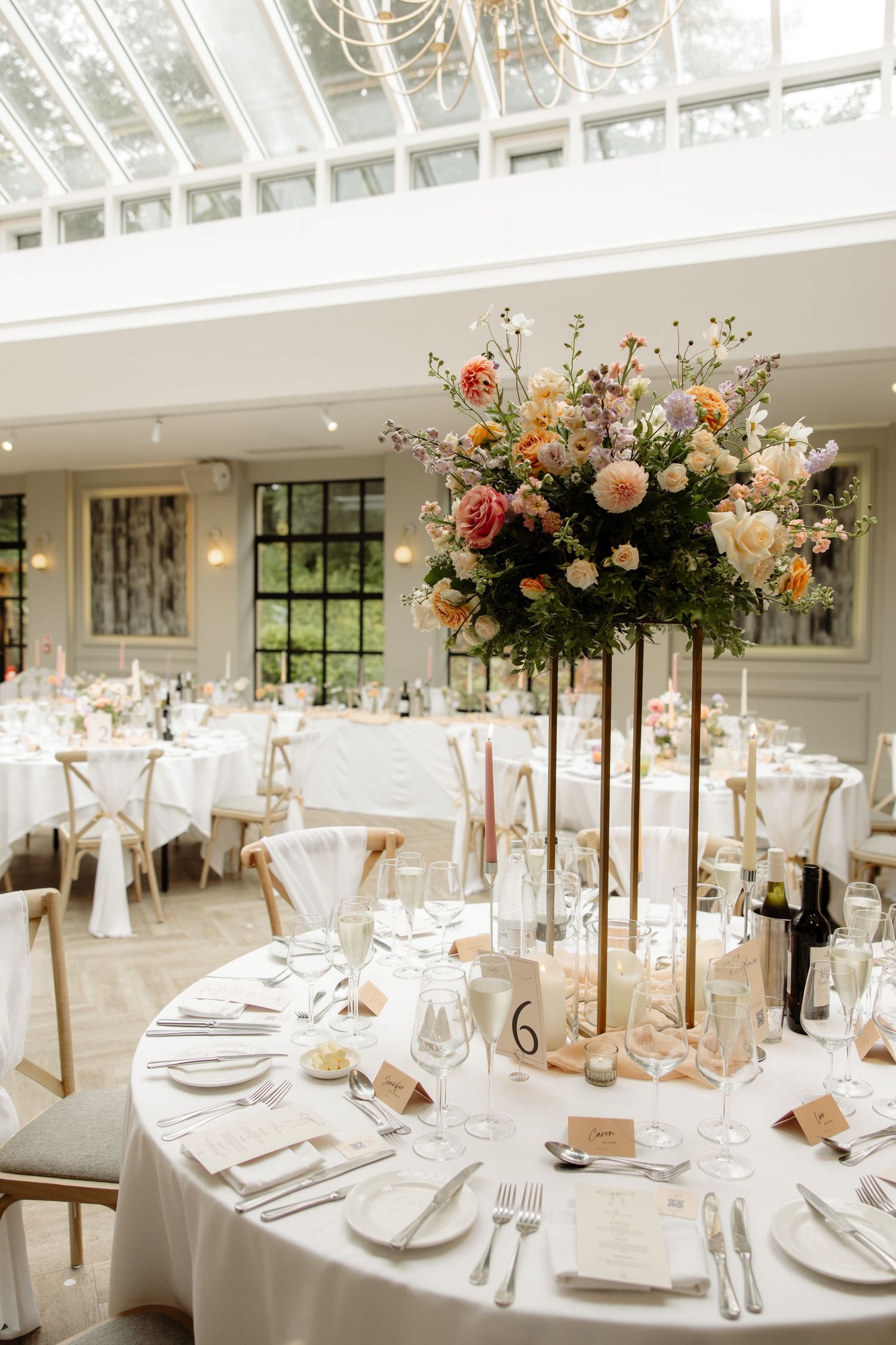 Elegant wedding reception tables set with white linens, tall floral centerpieces and golden candelabras in a light-filled venue.