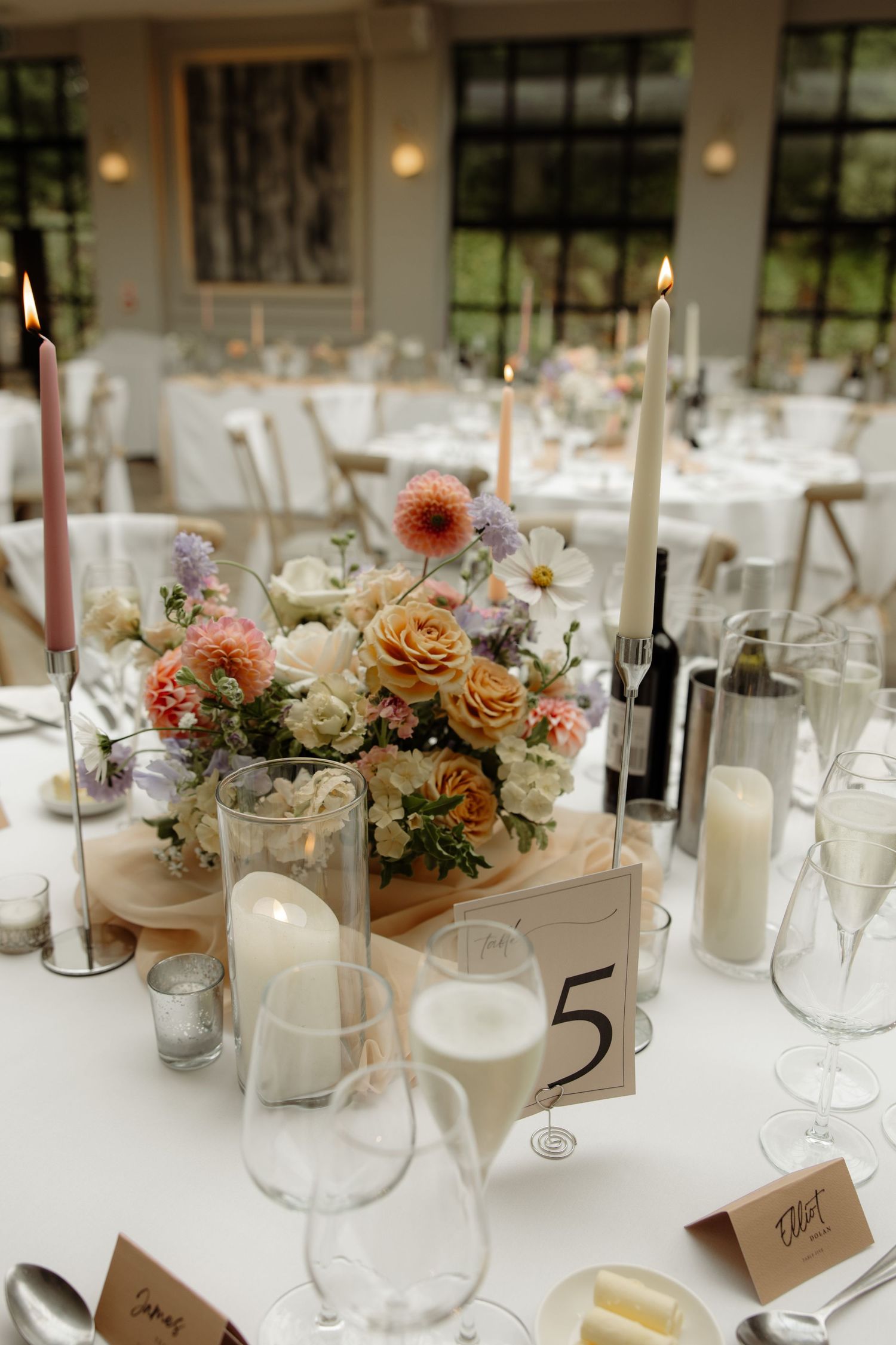 Elegant wedding reception table setting with white candles, peach and cream floral centerpieces, and wine glasses.