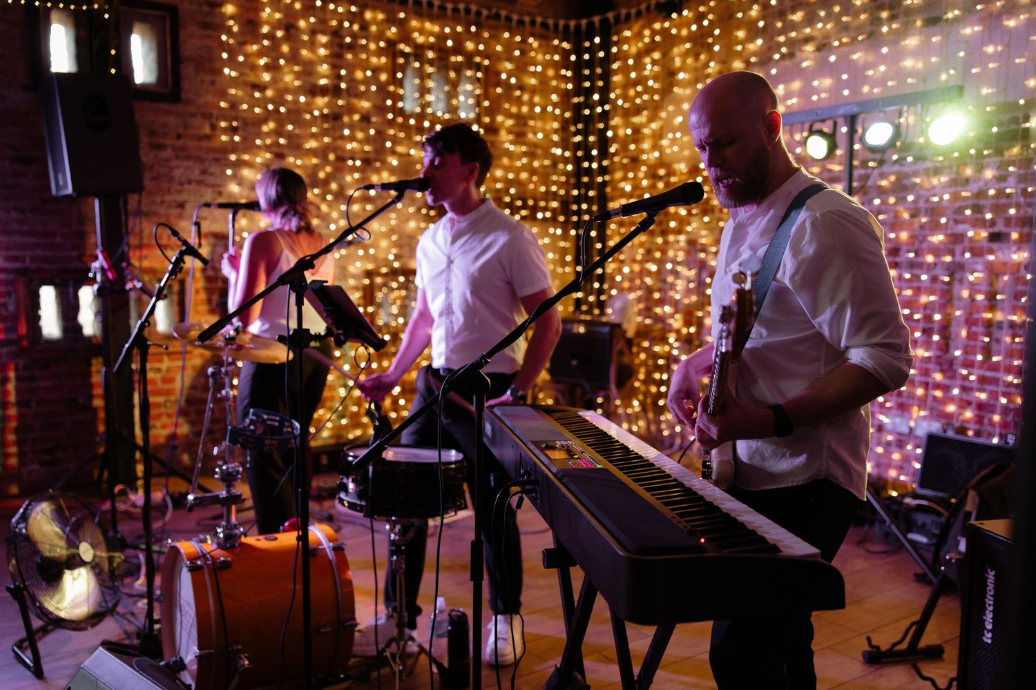 Musicians perform on stage with warm glowing string lights creating an intimate ambiance in the background.