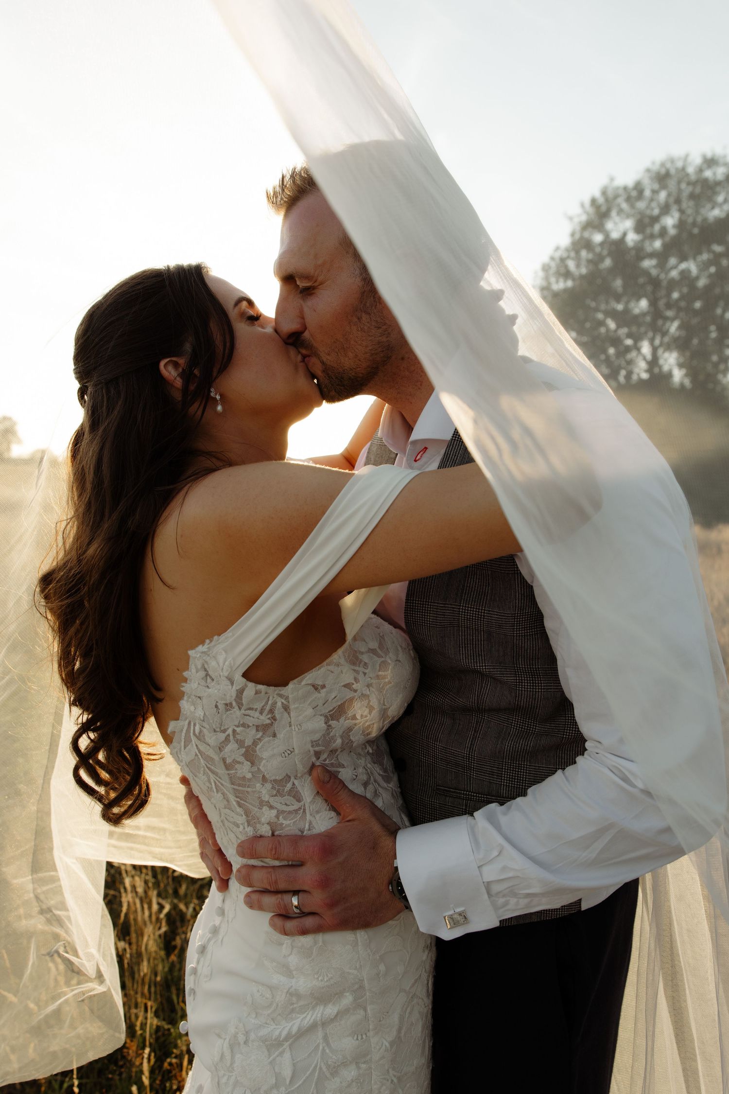 Romantic wedding couple embraces and kisses under flowing white veil during sunset in outdoor setting.