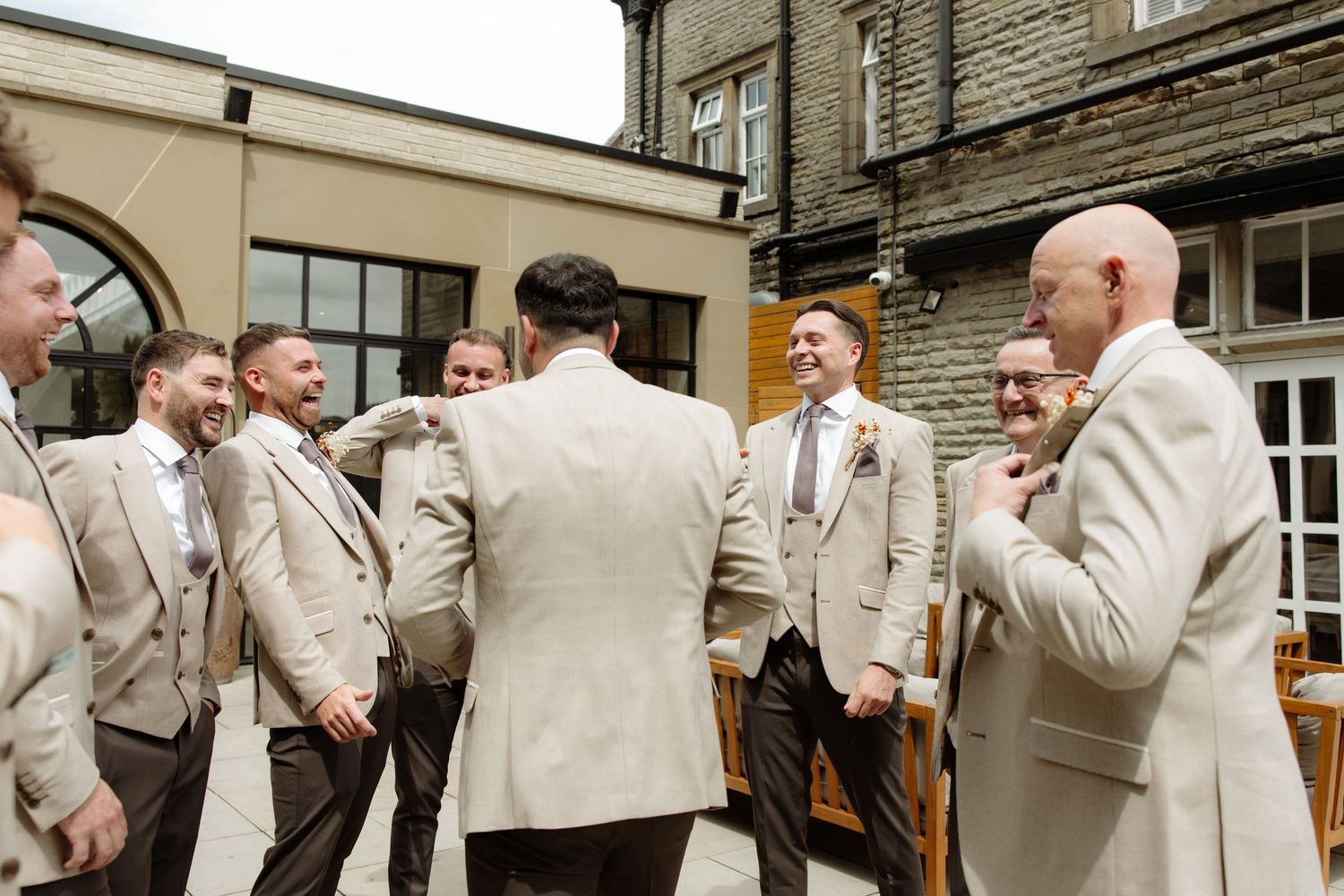 A group of groomsmen in beige suits gather and share laughs outside a stone building during a wedding celebration.