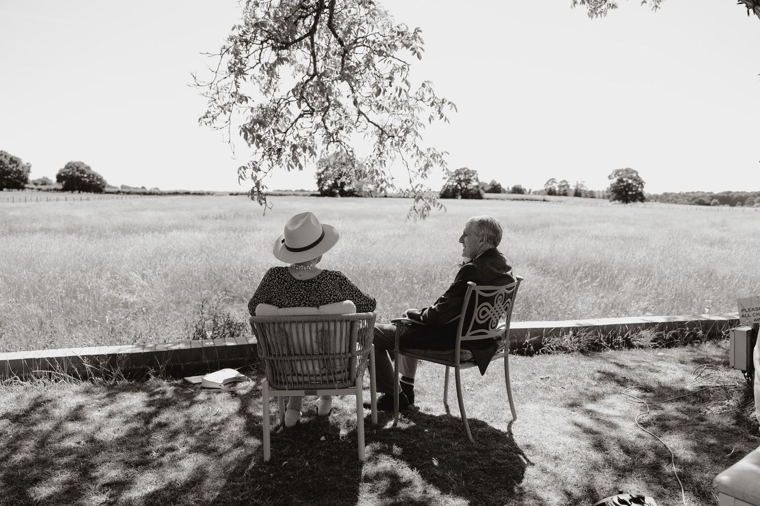 Two people sit on a bench overlooking a scenic countryside landscape on a sunny summer day.