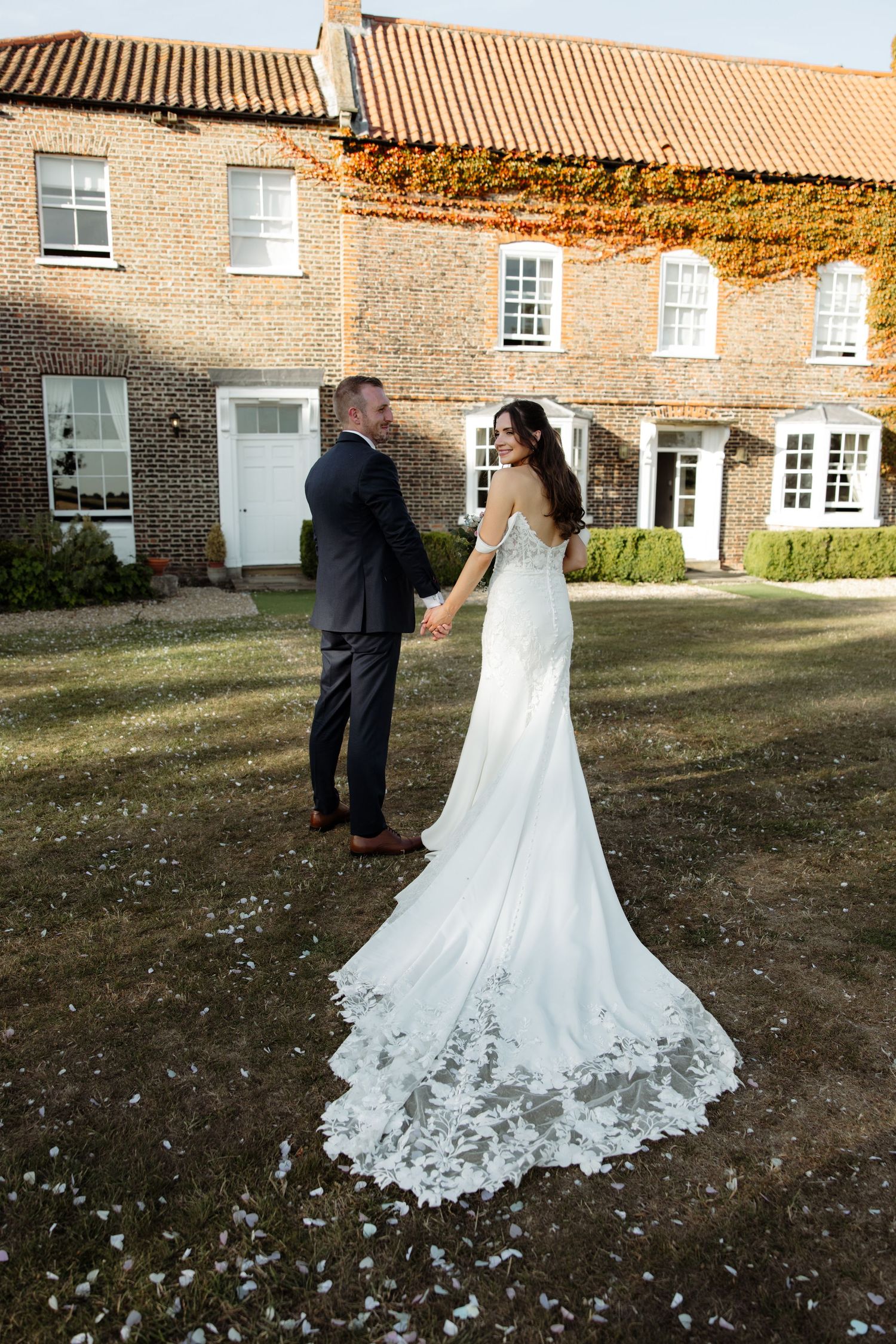 A romantic wedding couple walks together outside a rustic brick building with a beautiful lace-detailed wedding dress train.