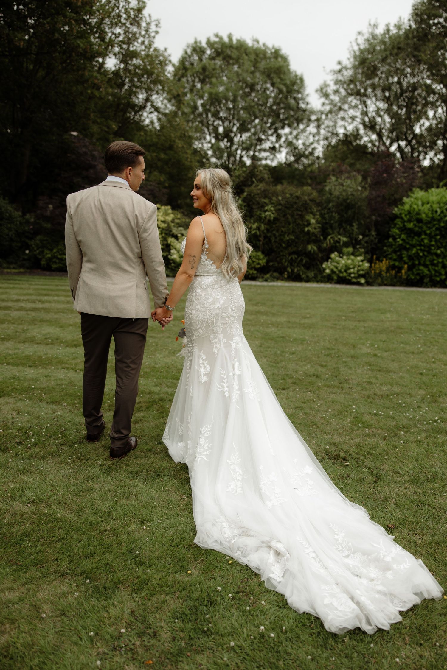 A couple walks across a lush green lawn with the bride's elegant white lace train trailing behind them in the grass.