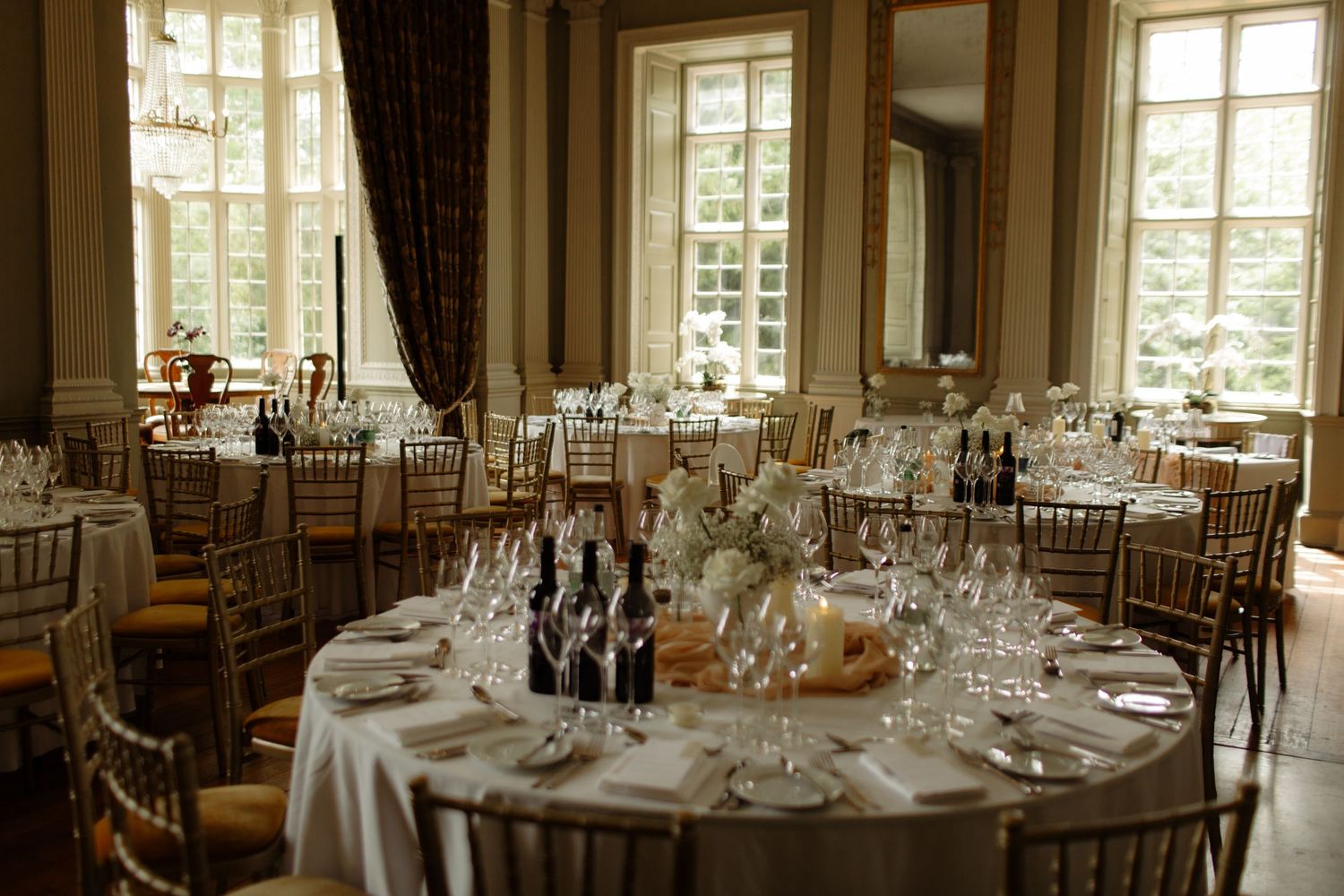 Elegant wedding reception tables set with wine bottles and glassware in a sunlit ballroom with tall windows.