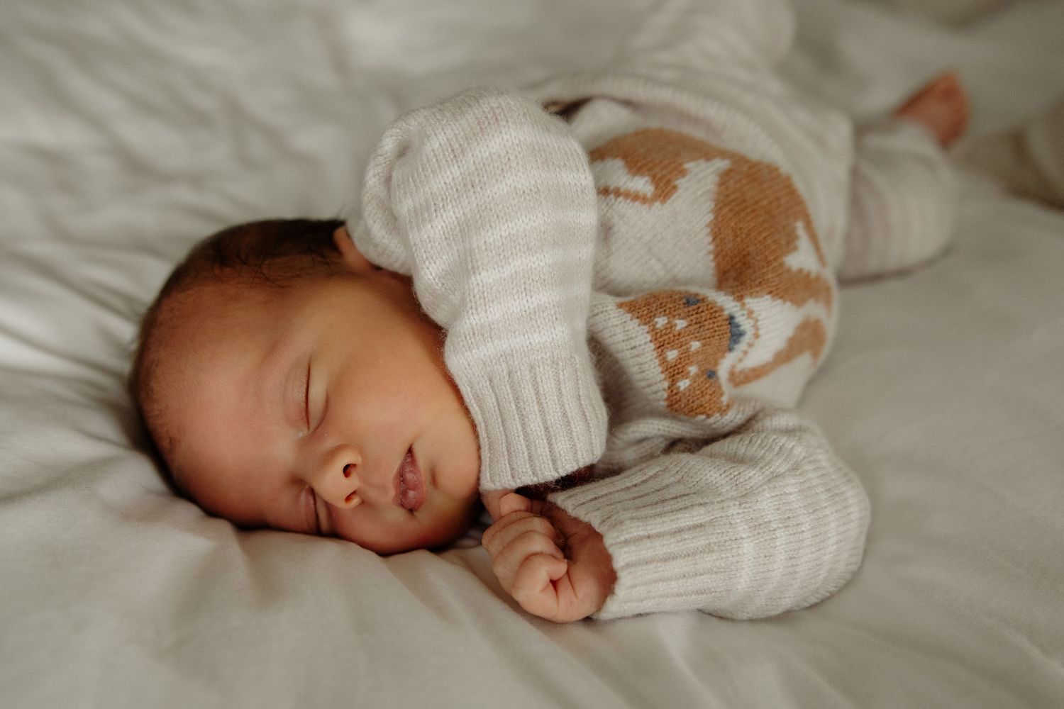 Newborn baby sleeping peacefully on white bedding wearing a cozy knit white and brown animal print onesie.