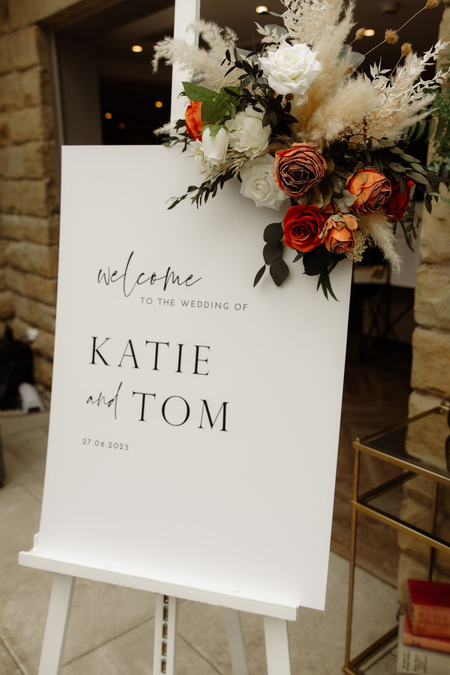 Elegant wedding welcome sign with 'Katie and Tom' text displayed on white cards with floral arrangements in the background.