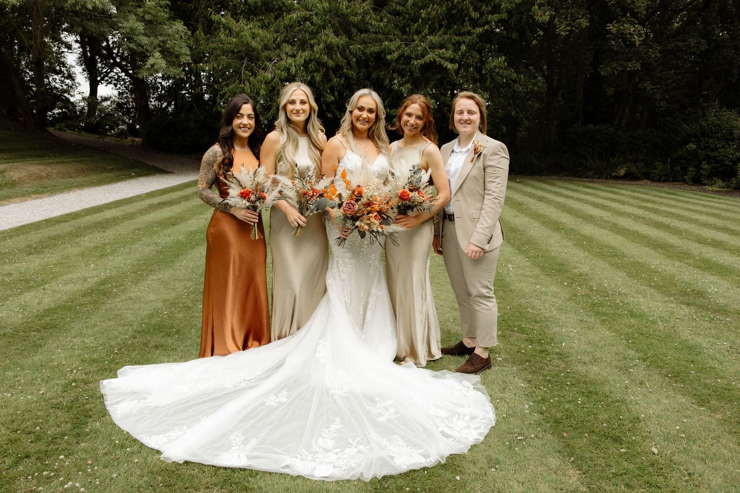 Wedding party stands together on a lush green lawn holding floral bouquets in autumn-toned dresses.