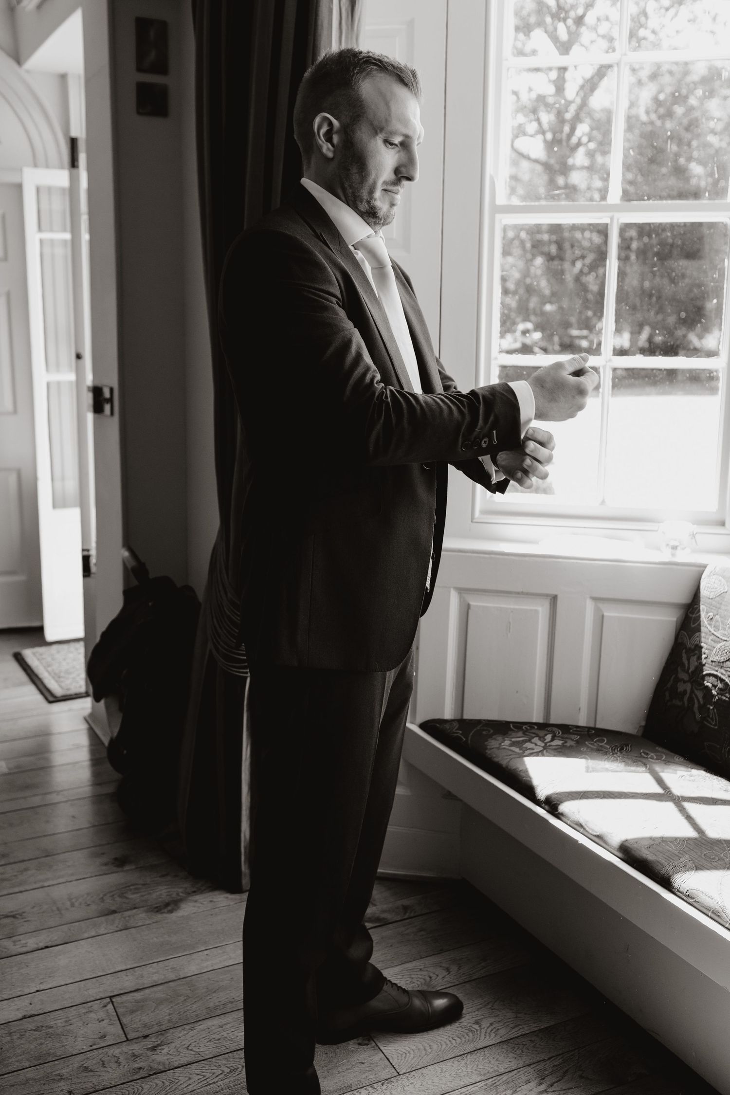 Black and white photo of a person in formal attire gazing pensively through a window in an elegant room.