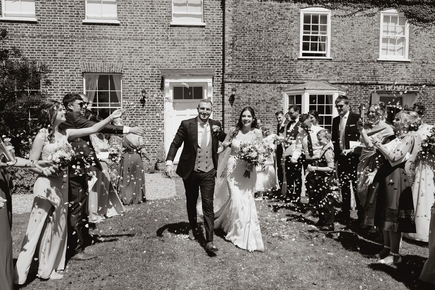 Wedding guests line up to throw confetti at a couple exiting a brick building in this black and white photograph.