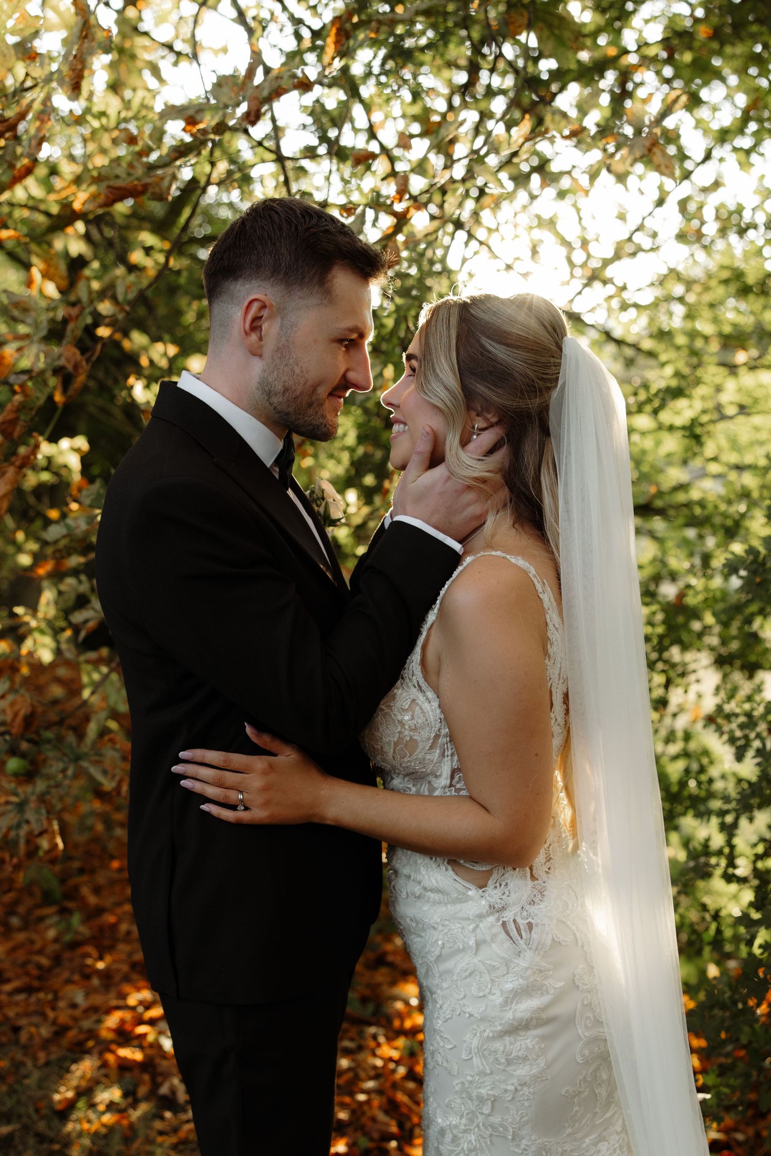 Romantic wedding couple embraces in golden sunlight filtering through autumn trees during outdoor photoshoot.