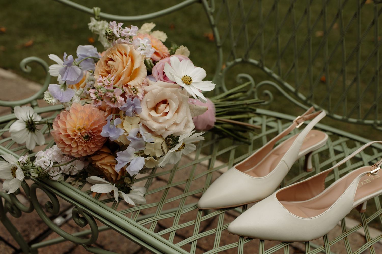 A delicate bridal bouquet with peach and blush roses sits next to ivory slingback heels on a vintage metal chair.