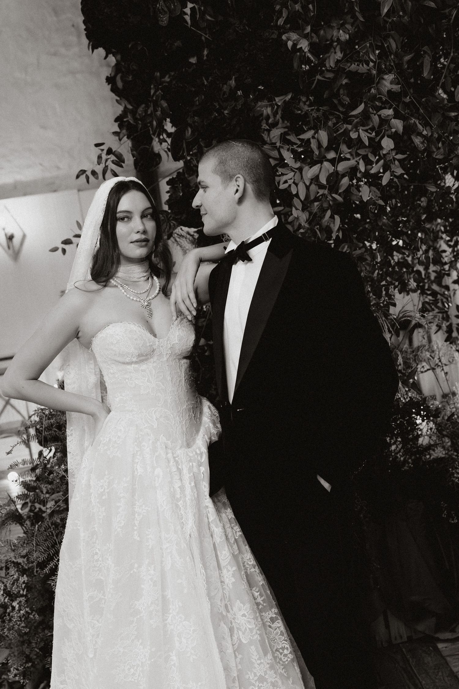 A bride in a white lace dress and groom in black tuxedo pose together against a leafy backdrop during their wedding.