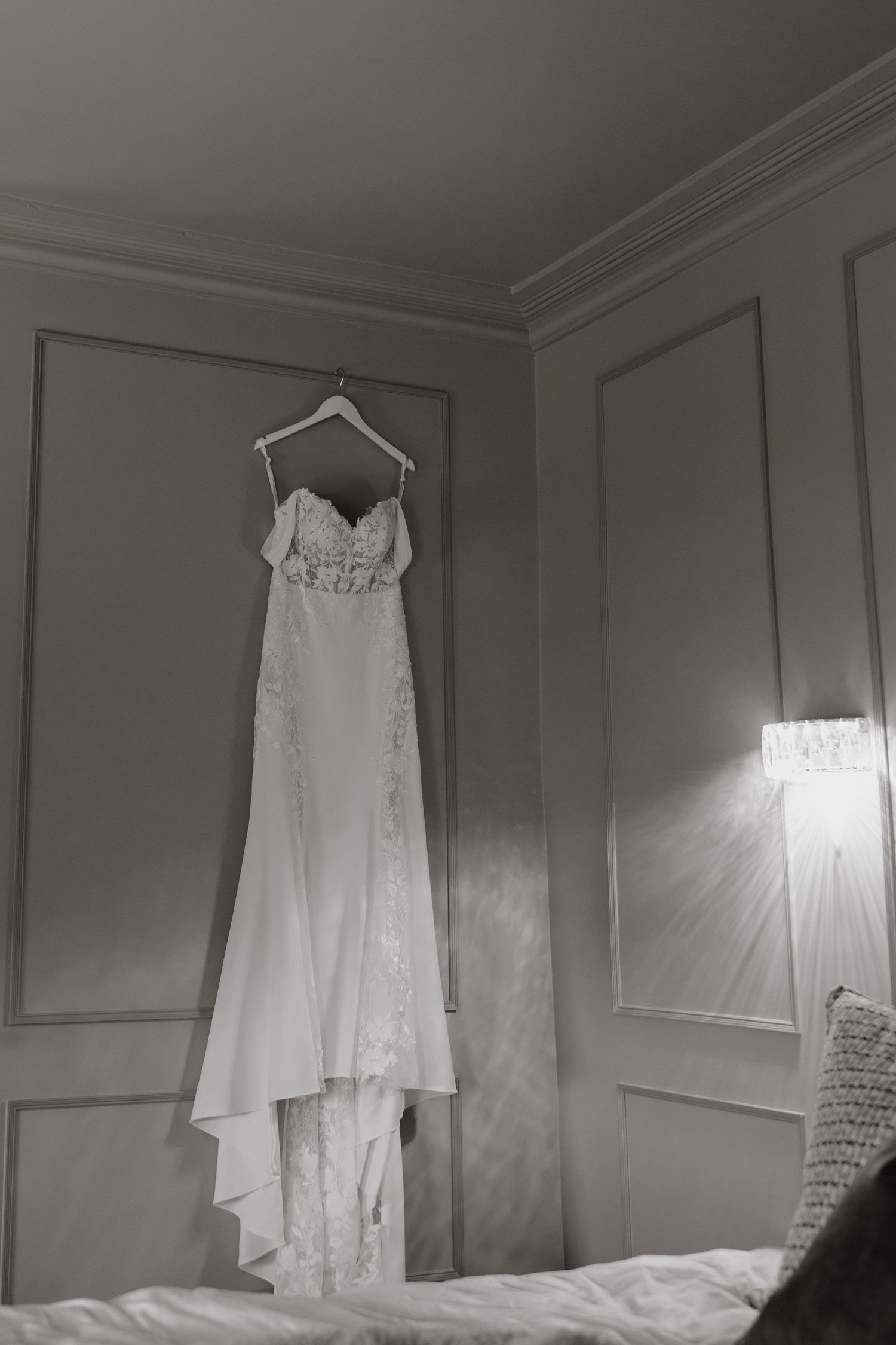 A white wedding dress with lace details hangs elegantly on a hanger against a light gray wall with classic molding.