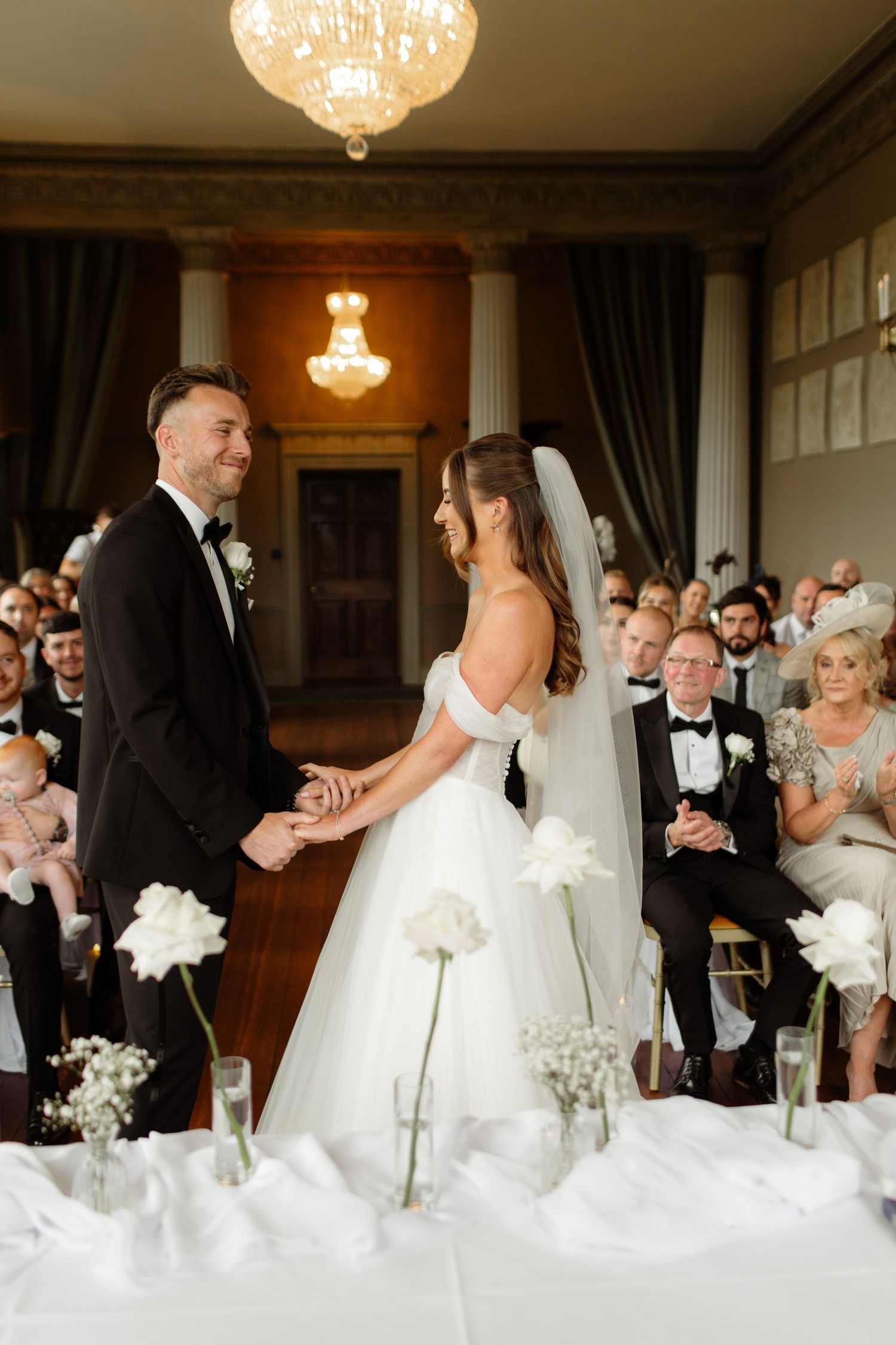 Elegant wedding reception in a ballroom with crystal chandeliers and white floral centerpieces on tables.