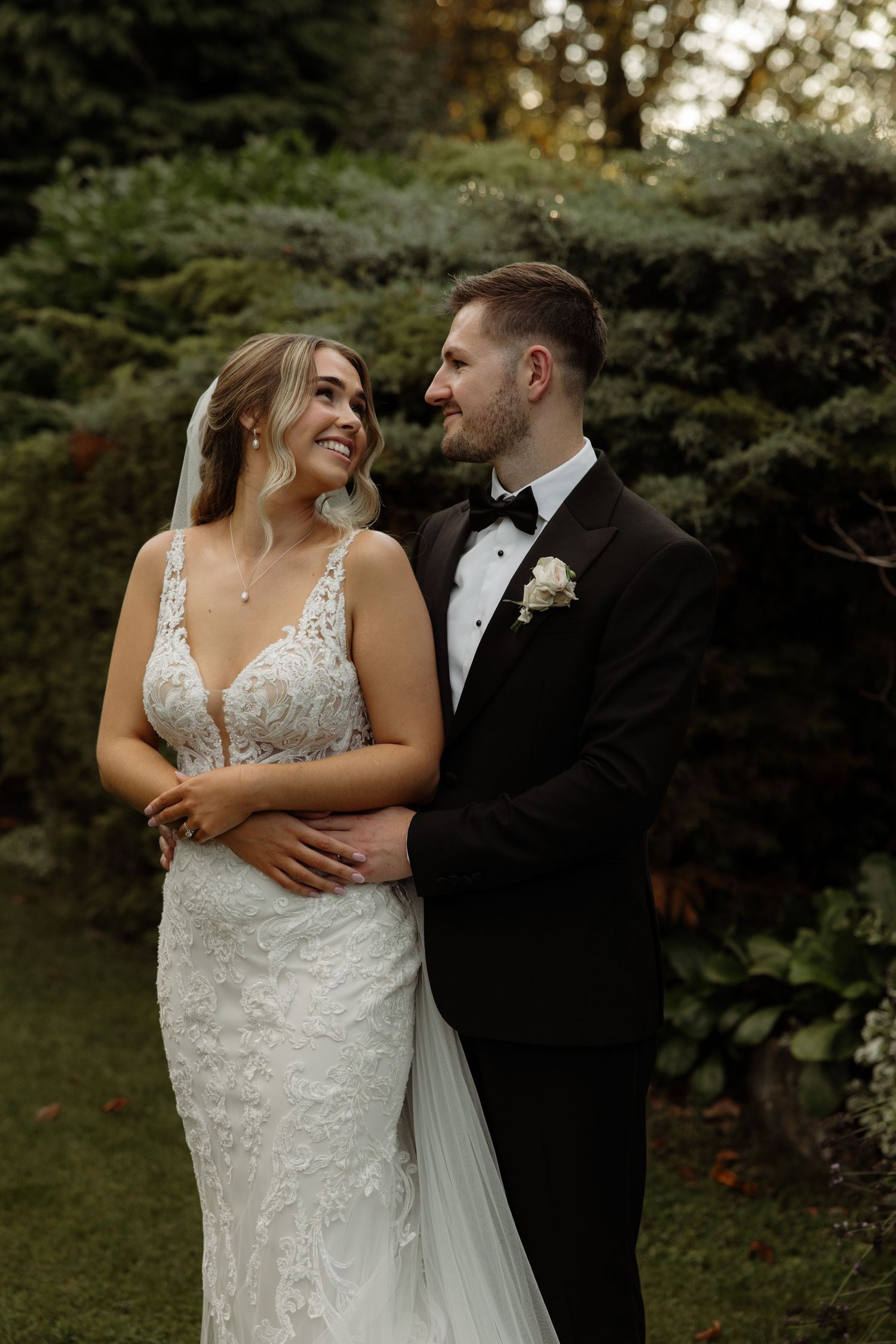 A couple shares intimate moments in their wedding attire against a backdrop of evergreen trees at dusk.