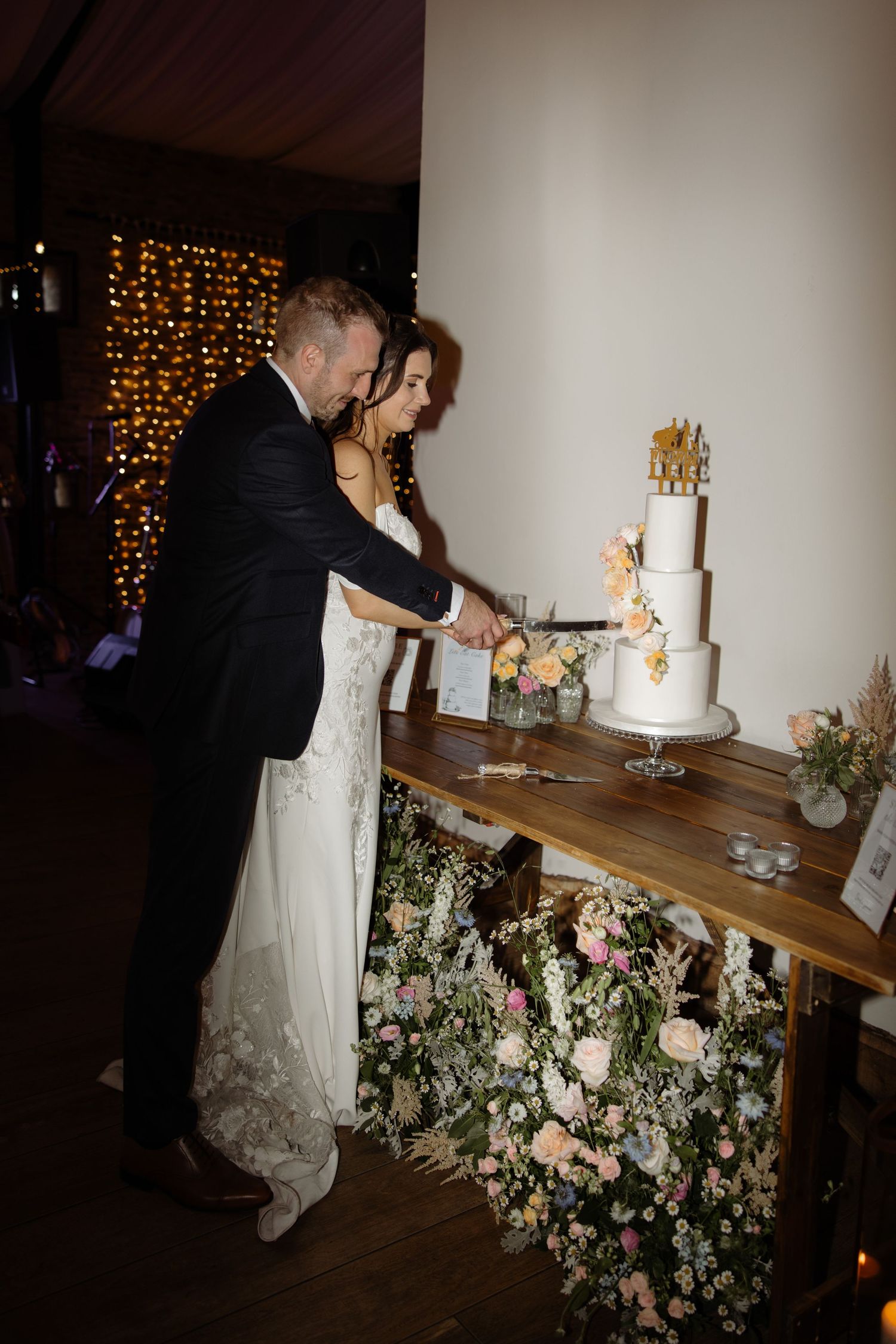 Couple cuts their elegant wedding cake decorated with flowers at a rustic wooden table with floral arrangements.