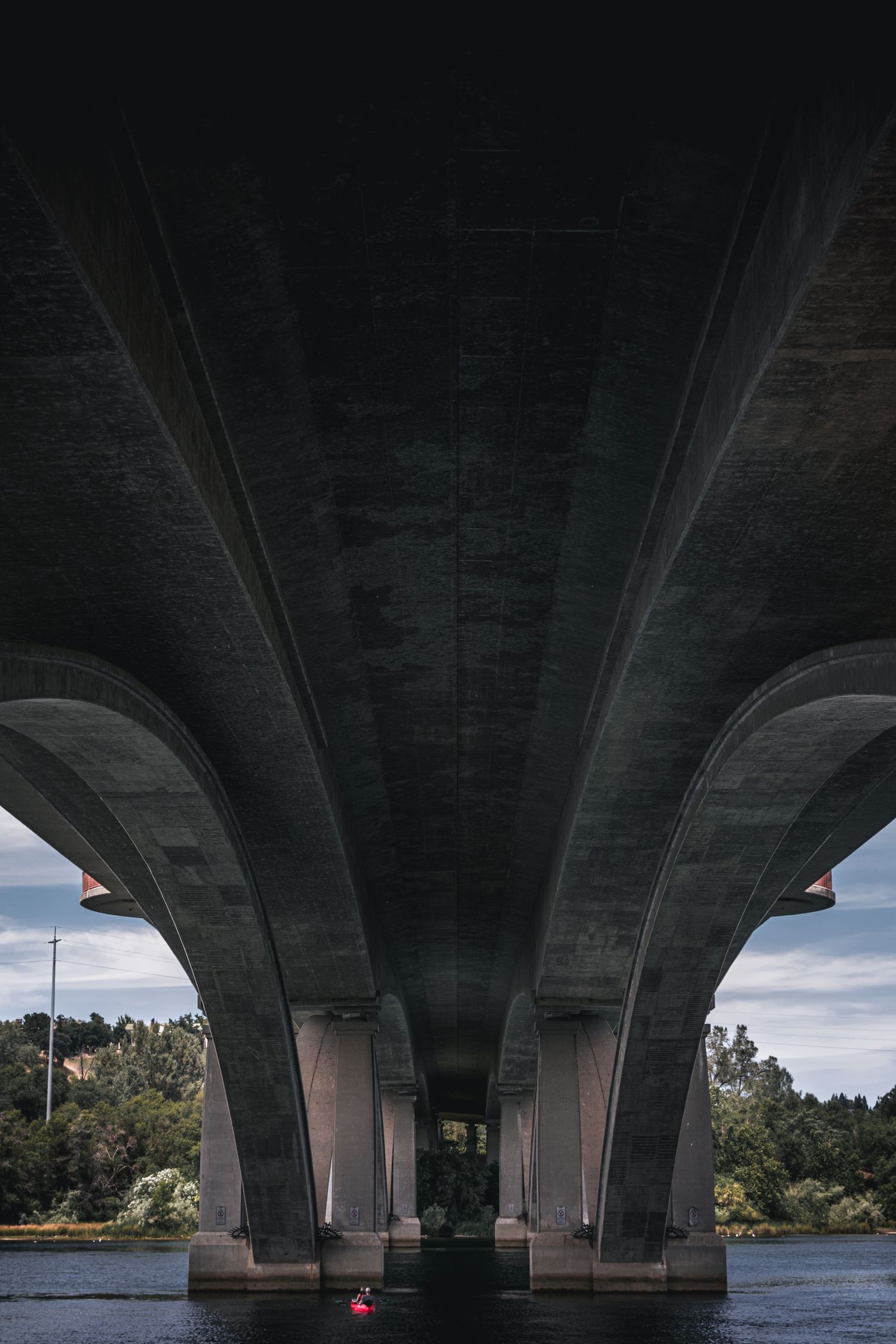 A low-angle shot looking up at the gray concrete arches of a large bridge over a body of water, with green trees in the background.