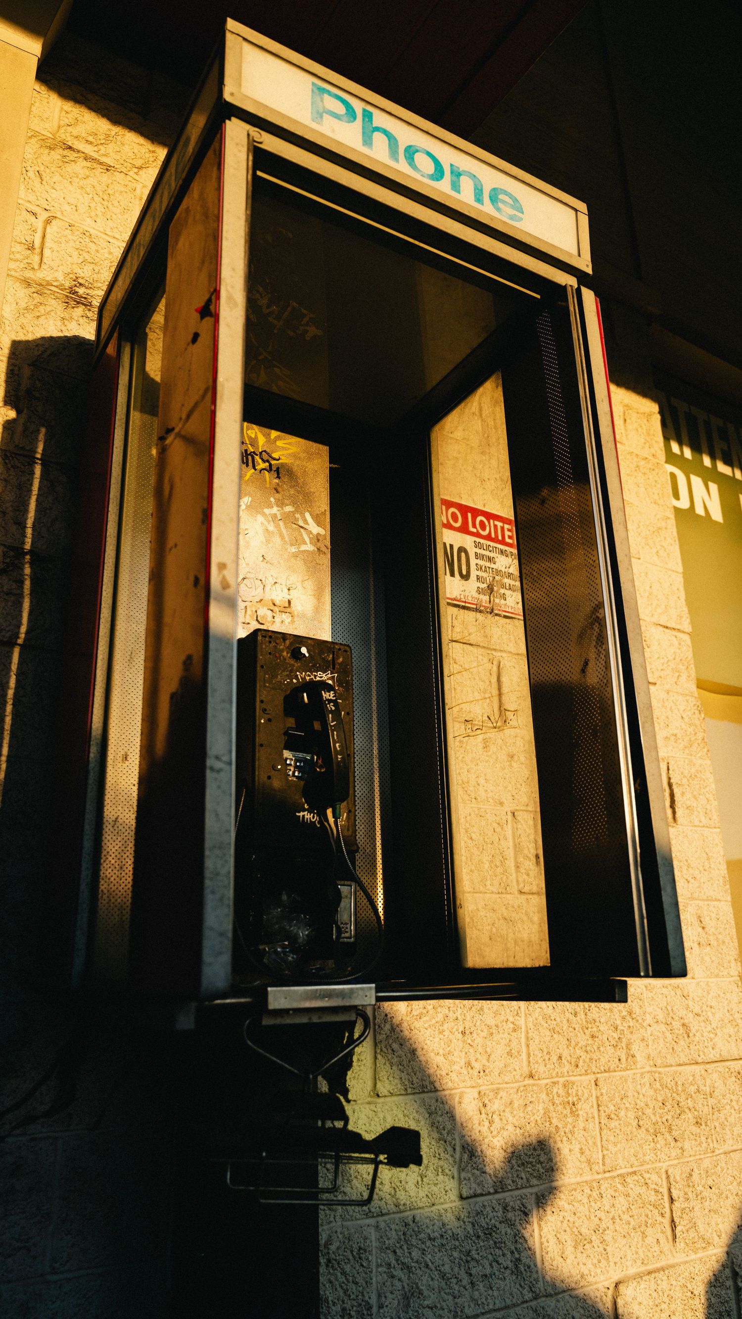 A vintage, empty public phone booth in an urban setting, illuminated by the warm, low-angle light of a sunset casting long shadows.