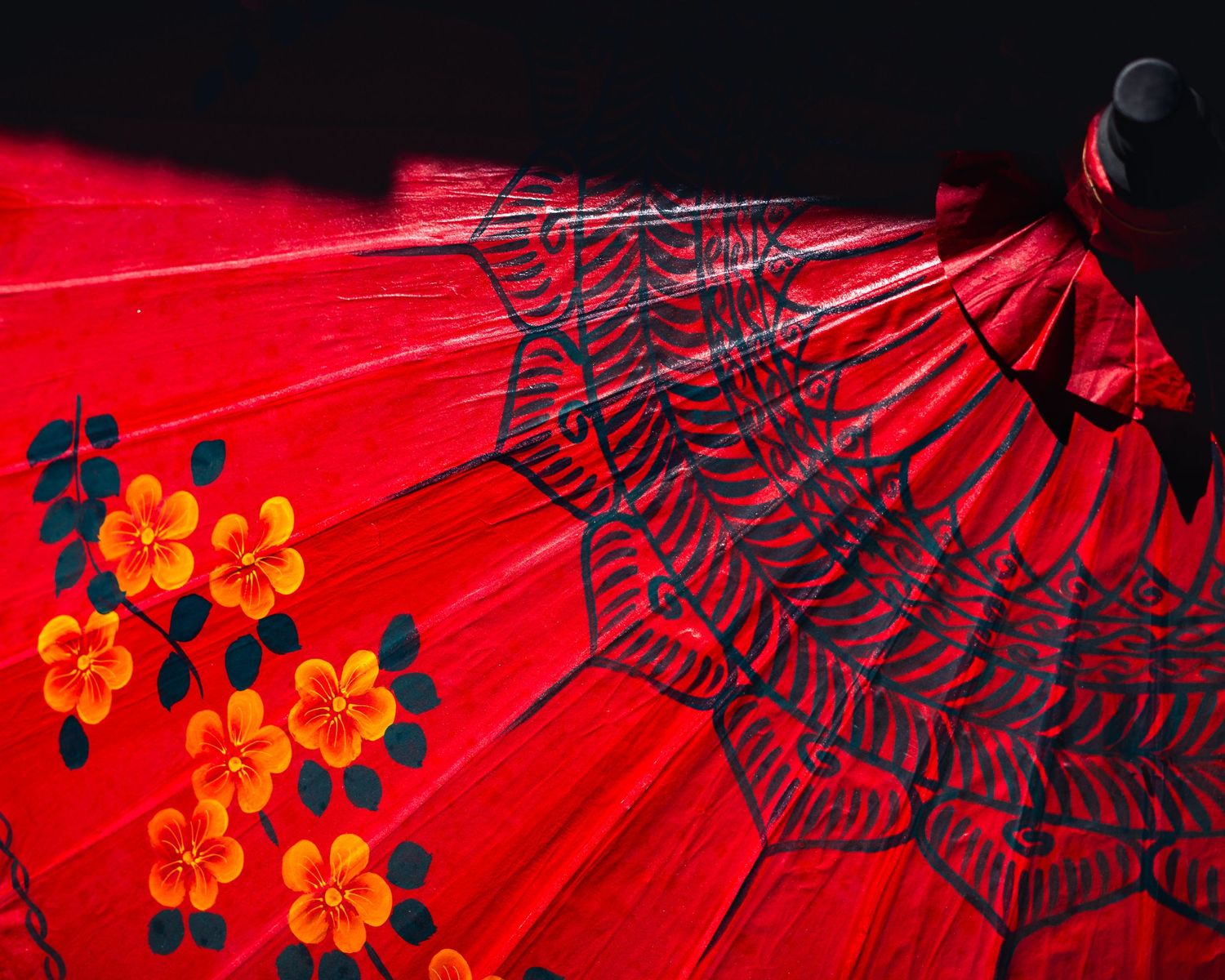 A close-up of a vibrant red umbrella, adorned with painted yellow and orange flowers and delicate dark blue patterns, partially shadowed.