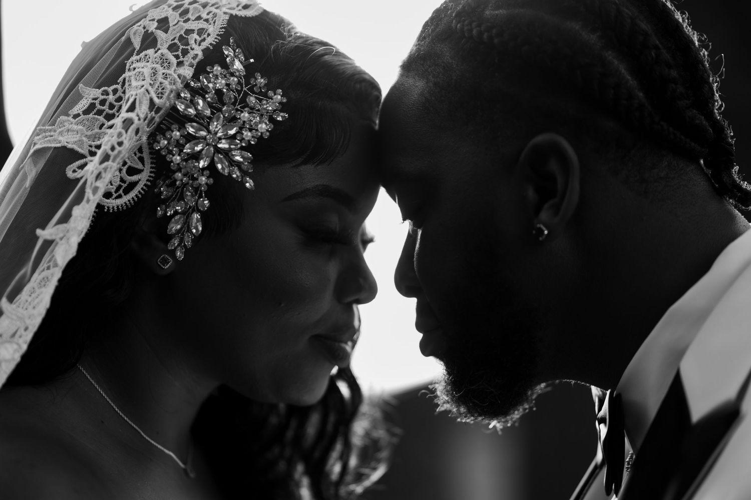 Black and white wedding photo of bride and groom touching foreheads, bride wearing lace veil and crystal headpiece.